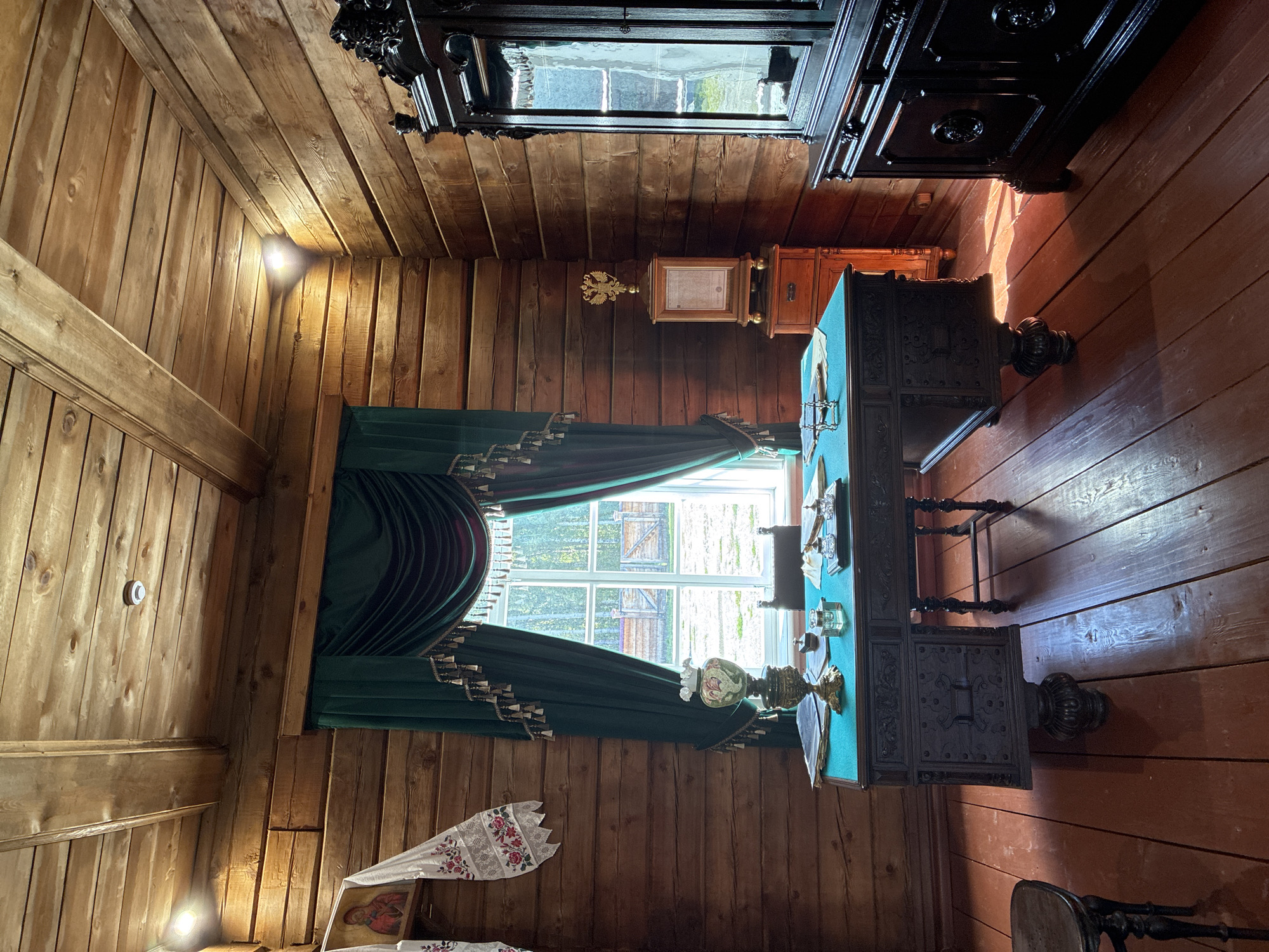 This is an interior shot of a room in a wooden log building, likely a museum exhibit, located in the Taltsy (Irkutsk) museum-reserve in Russia. The room is furnished as a study or office. The walls and ceiling are constructed of rough-hewn wooden logs. In the foreground, a large, dark, ornate wooden desk with carved details sits on a red wooden floor. The desk has a teal-colored surface with papers, a lamp with a decorative base and a white shade, inkwells, and other writing implements. To the left of the desk, a portion of a dark wooden chair is visible. Behind the desk, a large window is draped with heavy, dark green velvet curtains with tasseled trim. The window looks out onto a rural landscape with trees and a wooden fence. To the right of the desk, a dark wooden cabinet or armoire with mirrored doors and ornate carvings stands against the wall. Further to the right, a smaller wooden chest of drawers with a decorative crest or seal on top is visible. On the wall to the left of the window, a piece of embroidered white fabric with red floral patterns hangs, alongside a small icon of what appears to be a religious figure. Lighting appears to be provided by ceiling-mounted fixtures, one of which casts a distinct shadow. The overall impression is of a historical, well-preserved room from a past era.