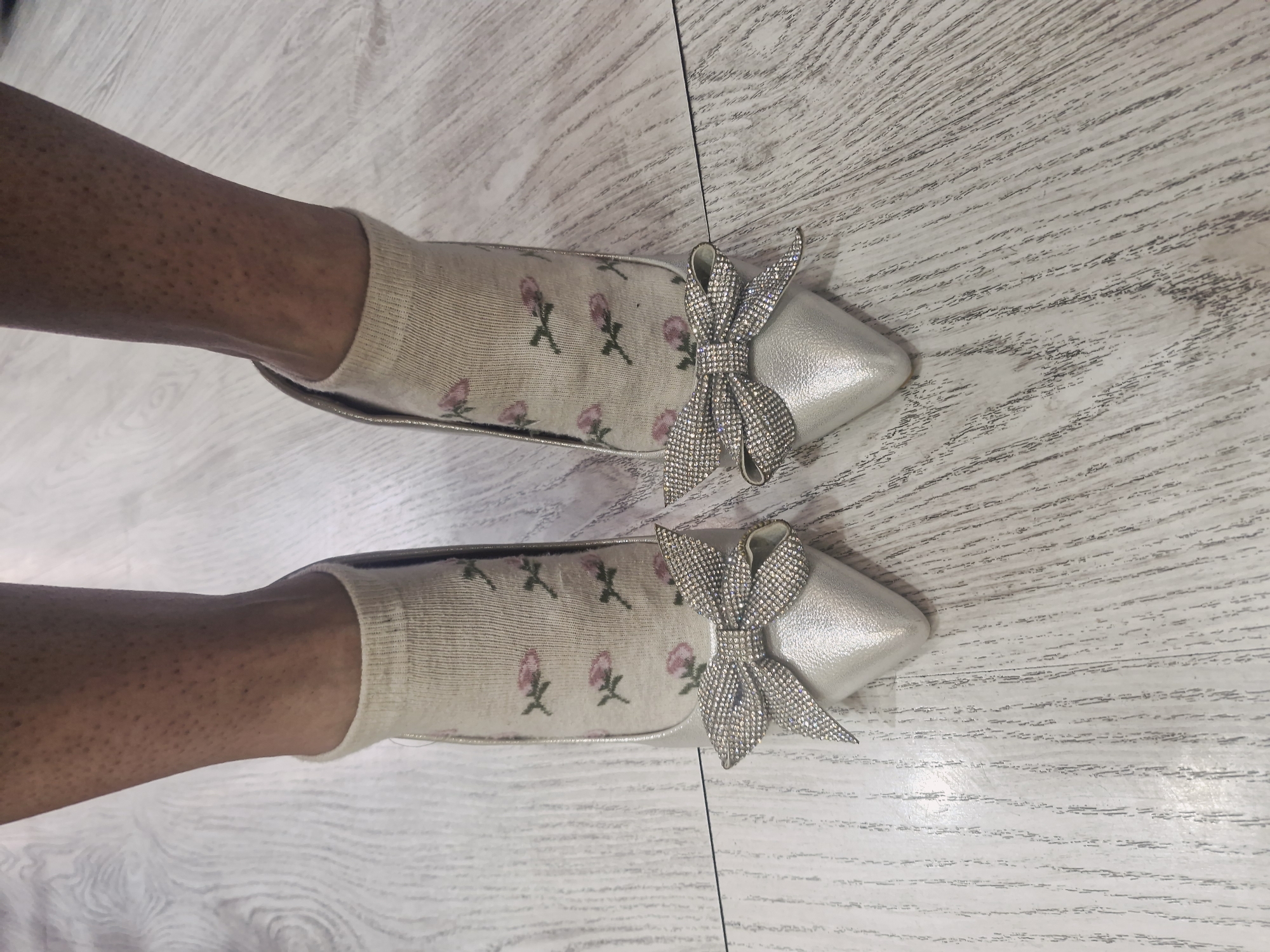 A top-down shot shows a pair of feet wearing beige socks with a pink floral pattern and silver, pointed-toe shoes adorned with glittery bows. They are positioned on a light-colored wooden floor with a striated pattern. There is no indication in the image that identifies the location as Plovdiv, Bulgaria.
