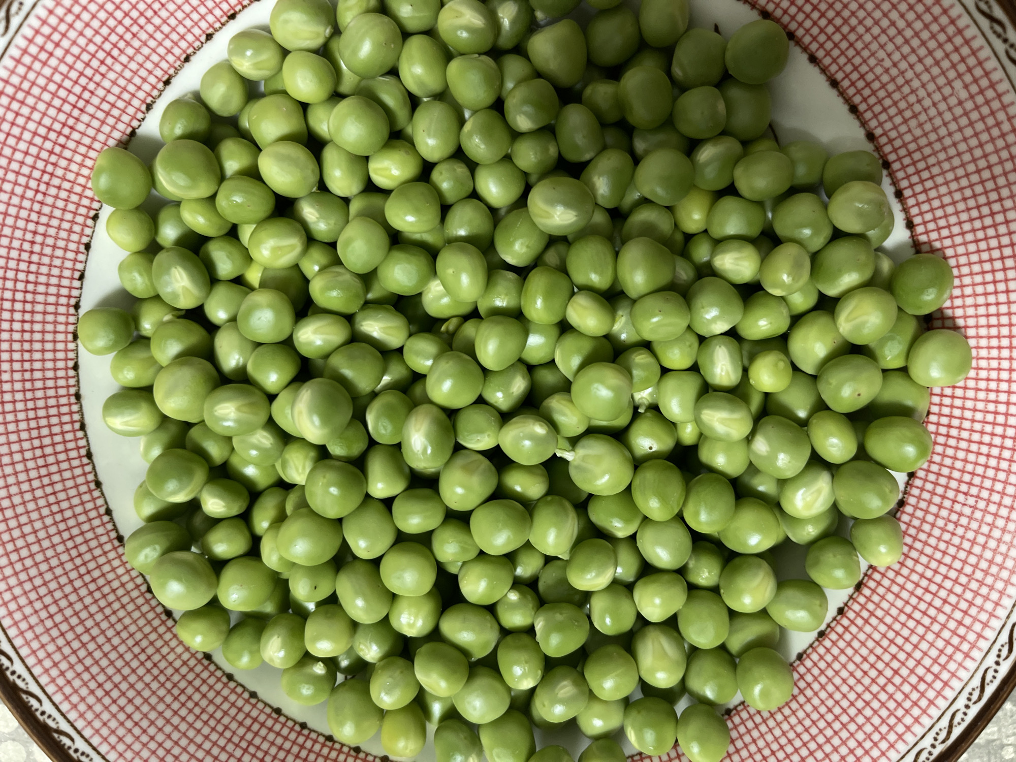 A close-up, overhead shot shows a large pile of fresh green peas filling a round, shallow bowl. The bowl has a white interior and a decorative rim featuring a repeating pattern of red squares outlined in brown. The peas are vibrant green, with a smooth, round surface, and some display subtle lighter-colored markings. The overall scene is bright and focuses entirely on the abundance of peas within the bowl. There is no visible indication of the location in the city of Средний, Russia.