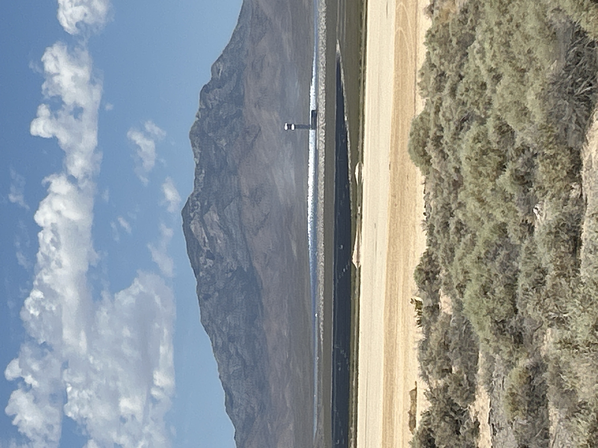 The image shows a vast solar power plant in the desert landscape near Primm, Nevada, USA. In the foreground, scrubby desert brush covers the sandy terrain. Further back, a large, dark expanse of solar panels stretches across the land, reflecting sunlight. In the middle ground, a tall, slender tower, characteristic of a solar thermal power plant, stands prominently. Behind the solar facility, a majestic mountain range rises, its slopes covered in muted tones of brown and grey. The sky above is a vibrant blue, dotted with fluffy white clouds. The overall scene depicts an arid, sun-drenched environment with a significant industrial presence.