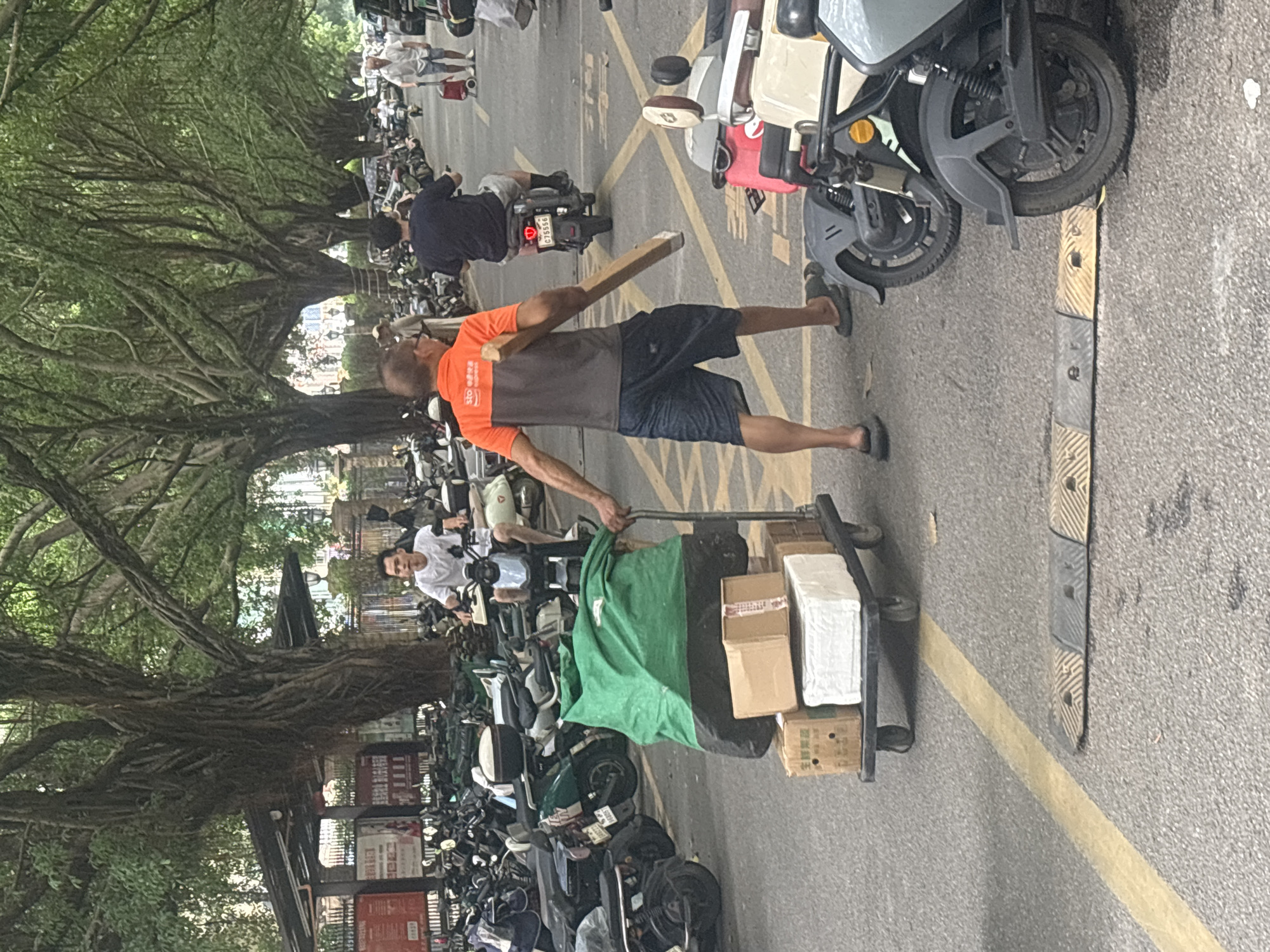 In Nanhai District, China, a man wearing an orange and grey polo shirt and dark shorts is pushing a cart laden with packages and a large green tarp. He holds a long, flat piece of wood in his right hand. The cart is positioned on a paved road, marked with yellow lines and a speed bump. Behind him, a man on a scooter with license plate C75556 is riding away. In the foreground, parts of parked electric scooters are visible. The background is filled with rows of parked scooters and motorcycles, shaded by large, mature trees with dense foliage. Other individuals are also visible in the distance, some on scooters and one walking with a red wheeled cart.