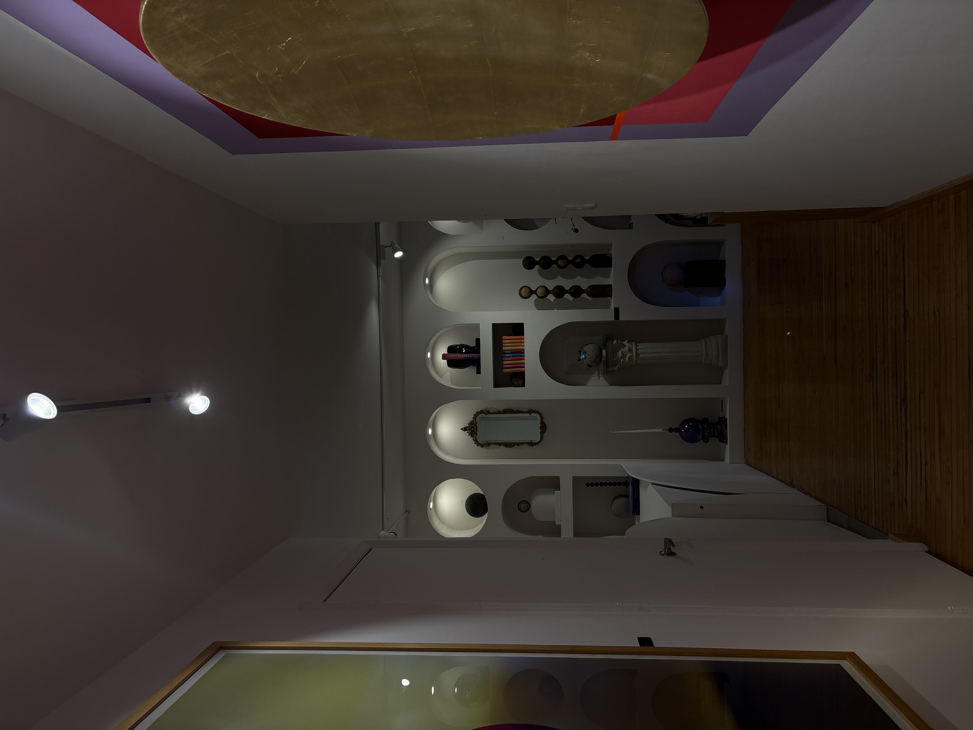 The image depicts an interior space in Mexico City, Mexico. The room features a series of arched niches built into a white wall, displaying decorative objects like wine bottles, books, a mirror, and sculptures. A wooden floor runs along the right side of the frame. Overhead, the ceiling showcases a decorative pattern with a gold circle, set against purple and red geometric shapes. Spotlights are mounted on the ceiling, illuminating the scene. The bottom of the frame shows a blurred, framed piece of artwork with a yellow border. The room appears to be an artfully decorated corridor.