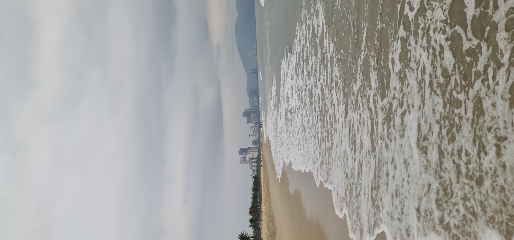 A ground-level, slightly-angled long shot shows a beach in Đà Nẵng, Vietnam. The shoreline runs horizontally in the midground from the center-left to the upper-right. The ocean's edge has white, foamy waves rolling in. The beach has tan sand.
In the midground, a line of palm trees stretches along the coast. Beyond the trees, a row of high-rise buildings forms the city skyline. In the distance, indistinct mountains fade into the hazy sky.
The sky is cloudy and overcast.