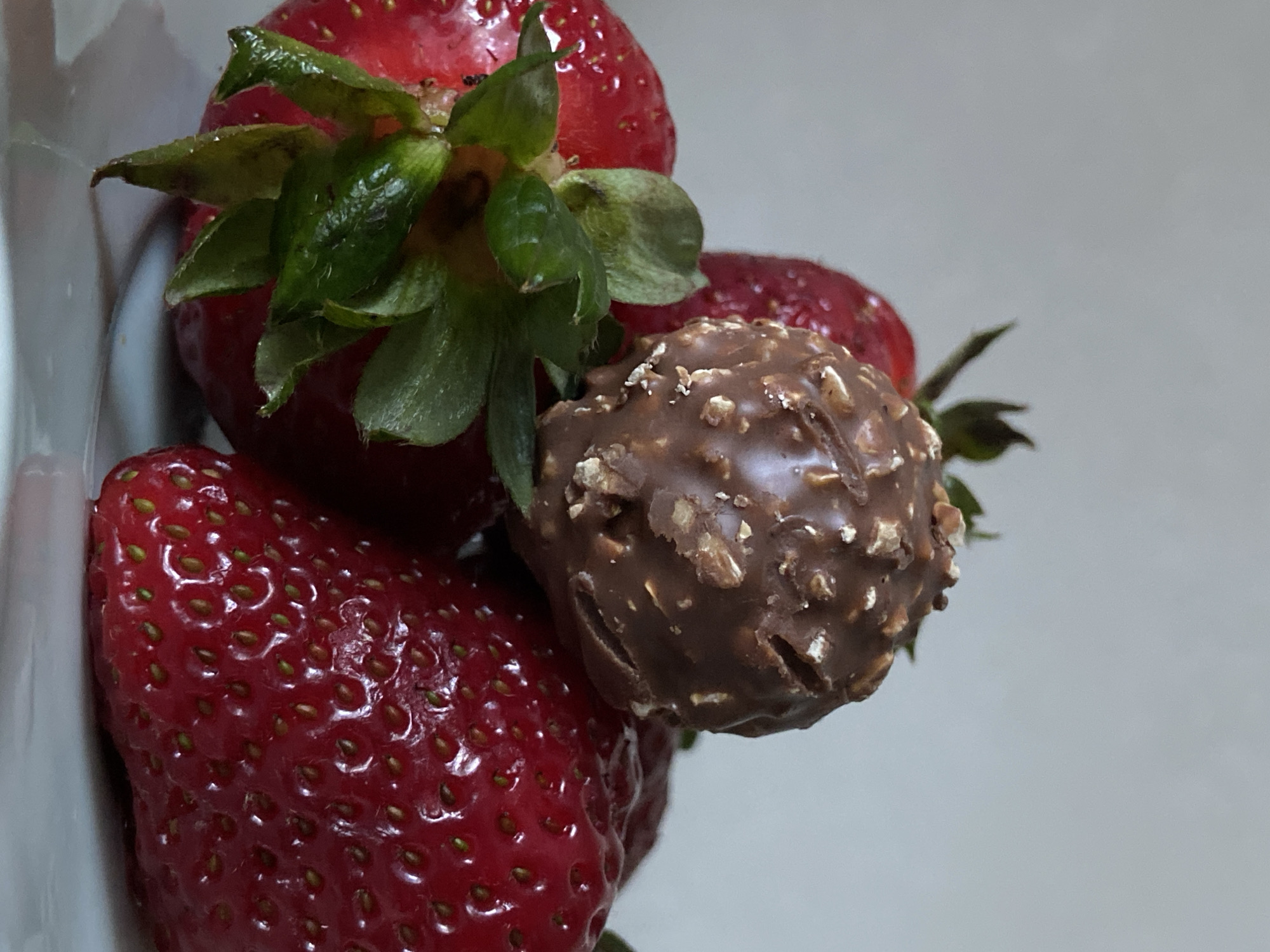 A close-up, slightly rotated shot captures a grouping of red strawberries and a chocolate candy against a plain, light background. Several vibrant red strawberries are visible, with prominent green leafy caps on the ones in the upper left and mid-right. The large strawberry in the lower-left foreground displays its textured surface and visible seeds. Nestled among the strawberries, primarily in the center-right, is a spherical chocolate candy, which is dark brown and covered in small, lighter-colored nut pieces, suggesting a crunchy coating. The items are resting on a white or light-colored surface, possibly a bowl or plate, with the background being a soft, light grey, offering a clean and simple setting. There are no people or discernible activities taking place. The lighting is bright and even, highlighting the details and textures of the fruit and the chocolate. No text is visible in the image.