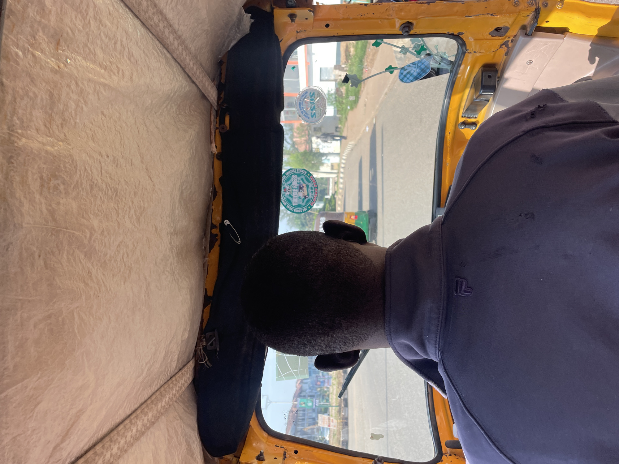 The image shows the interior of a yellow auto-rickshaw, likely a commercial taxi, with the camera positioned behind the driver, looking forward. The driver, a Black male with short hair, is wearing a dark purple collared jacket with a small, subtle logo on the left side of the back. The interior of the auto-rickshaw appears to be made of light-colored, textured material on the roof and black padding along the top of the windshield.
The windshield offers a view of a street scene in what is identified as Jos, Nigeria. The road is paved and appears to be a busy thoroughfare. There are other vehicles visible, including a green auto-rickshaw in the left lane and a person walking on the sidewalk in the distance. Buildings and trees line the street, suggesting an urban environment. Two circular stickers are affixed to the inside of the windshield. The sticker on the left has text and an image of auto-rickshaws, reading "SOUTH LOCAL GOVERNMENT TRICYCLE RIDERS PLATEAU STATE." The sticker on the right is partially obscured but appears to be another official-looking emblem. The overall lighting suggests it is daytime.