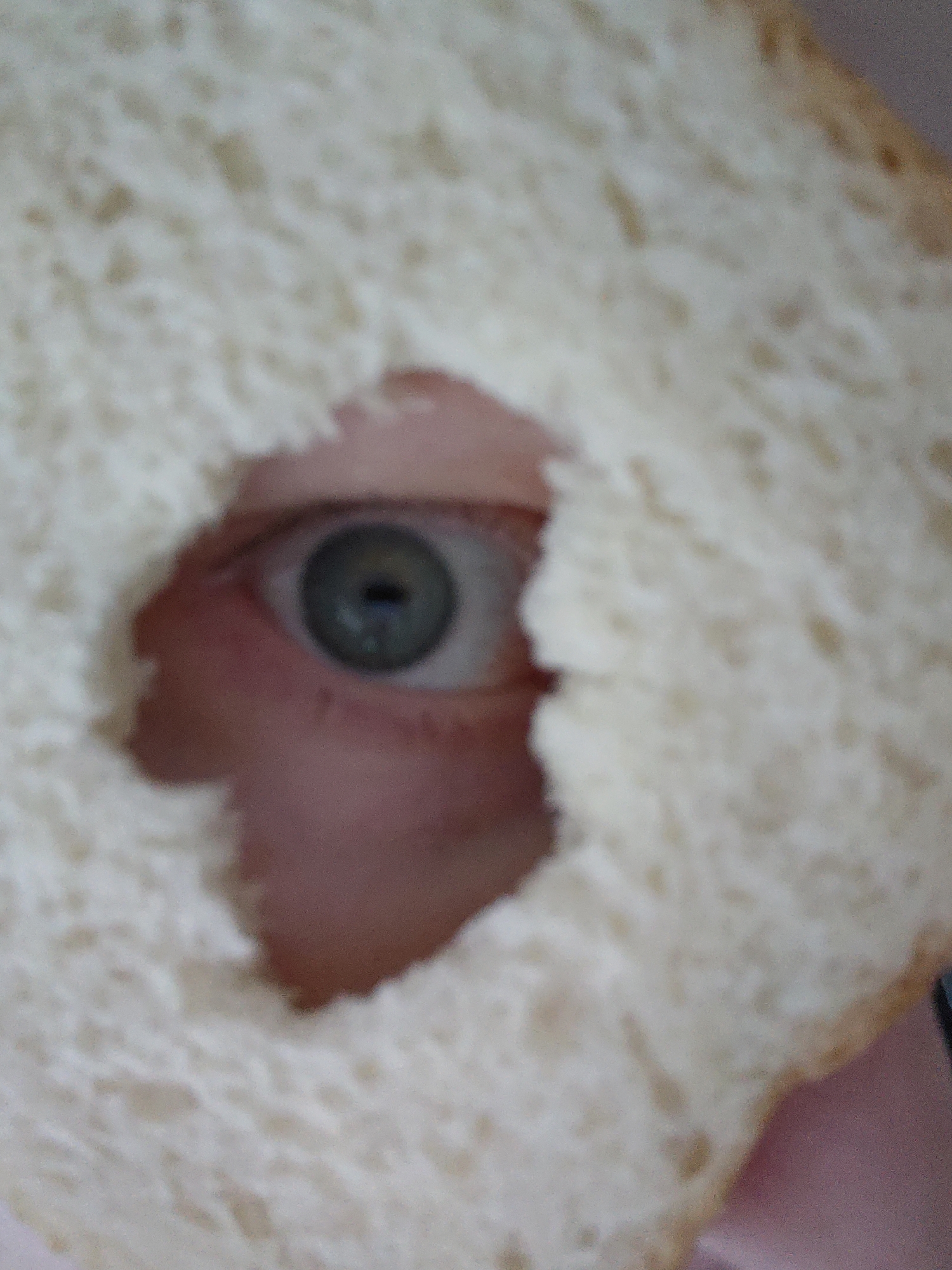 A close-up, slightly blurred image shows a human eye peering through a hole in a slice of white bread. The eye is light-colored with a dark pupil. The surrounding skin is pinkish. The bread is soft and white with a slightly darker crust visible. The photo is taken at close range, filling the frame with the bread and eye. There is no information to suggest this image was taken in Markovskii, Russia.