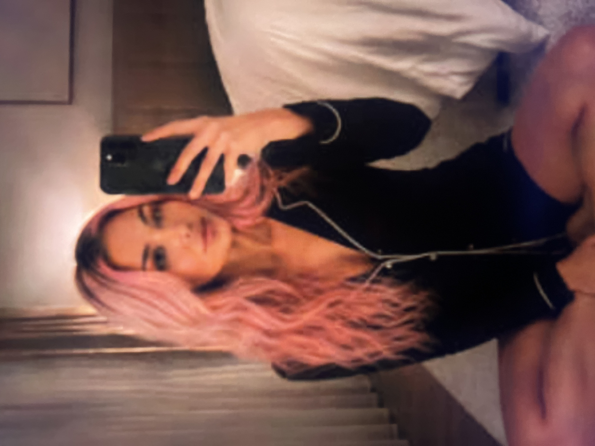 An individual with long, wavy, pink-dyed hair is taking a selfie in a dimly lit room. They are wearing black pajamas with white piping. The individual is sitting cross-legged on the floor, with their legs crossed in front of them. A white duvet is visible on a bed in the background, along with a headboard and a framed picture on the wall. A sheer curtain is on the left side of the image. The lighting suggests it may be nighttime or the room is dimly lit.