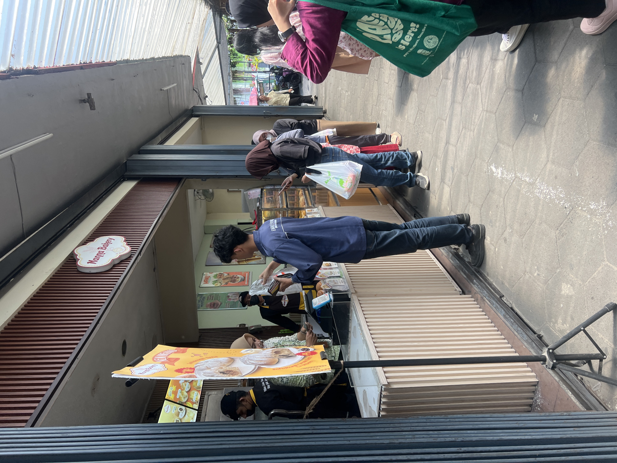 The image depicts a street-level view of a Manna Bakery in Yogyakarta, Indonesia. A queue of people is lined up outside the establishment, with several individuals interacting with staff members at a counter. The bakery features a wooden facade and a sign with its name. A prominent yellow promotional banner displaying images of pastries is positioned in the foreground, supported by a tripod. The scene is outdoors, with people dressed casually in attire suitable for the climate. The ground is paved with hexagonal tiles. The overall ambiance suggests a typical day at a popular food vendor in a city setting.
