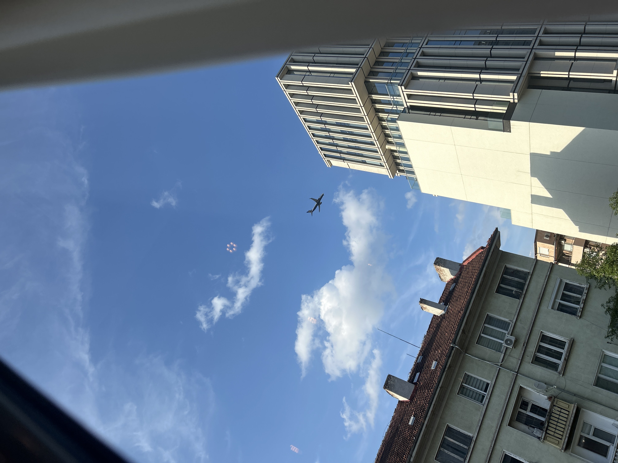 The image captures an urban landscape under a clear blue sky with scattered white clouds, seen from an elevated perspective, likely through a window. A dark silhouette of an airplane is visible in the upper-middle left portion of the sky, appearing to be in ascent or descent. Several faint, circular, multi-ringed lens flares or reflections are present in the sky, notably in the upper right and upper left quadrants.
On the left side of the frame, an older, multi-story residential building occupies the lower-left. It features a light green-grey stucco facade, numerous rectangular windows, some with white frames and external shutters, and at least one small balcony with an ornate metal railing. The building is topped with a steeply pitched, reddish-brown tiled roof from which several rectangular chimneys emerge. An antenna or thin wire extends upwards from the roof.
Dominating the right side of the frame is a modern, high-rise building constructed with light-colored (cream or white) materials, featuring distinctive vertical architectural elements and large, dark-tinted glass window sections. The building exhibits multiple setbacks and terraces with clear glass railings. Its upper section displays prominent vertical fins. This modern building's architecture is consistent with the NV Tower in Sofia, Bulgaria.
Partial obstructions, appearing as a dark, angled frame element in the upper left corner and a wider, light-colored vertical element on the far right, suggest the photograph was taken from within an interior space, likely looking out a window. The combination of traditional and contemporary architecture is characteristic of the evolving urban fabric of Sofia, Bulgaria.
