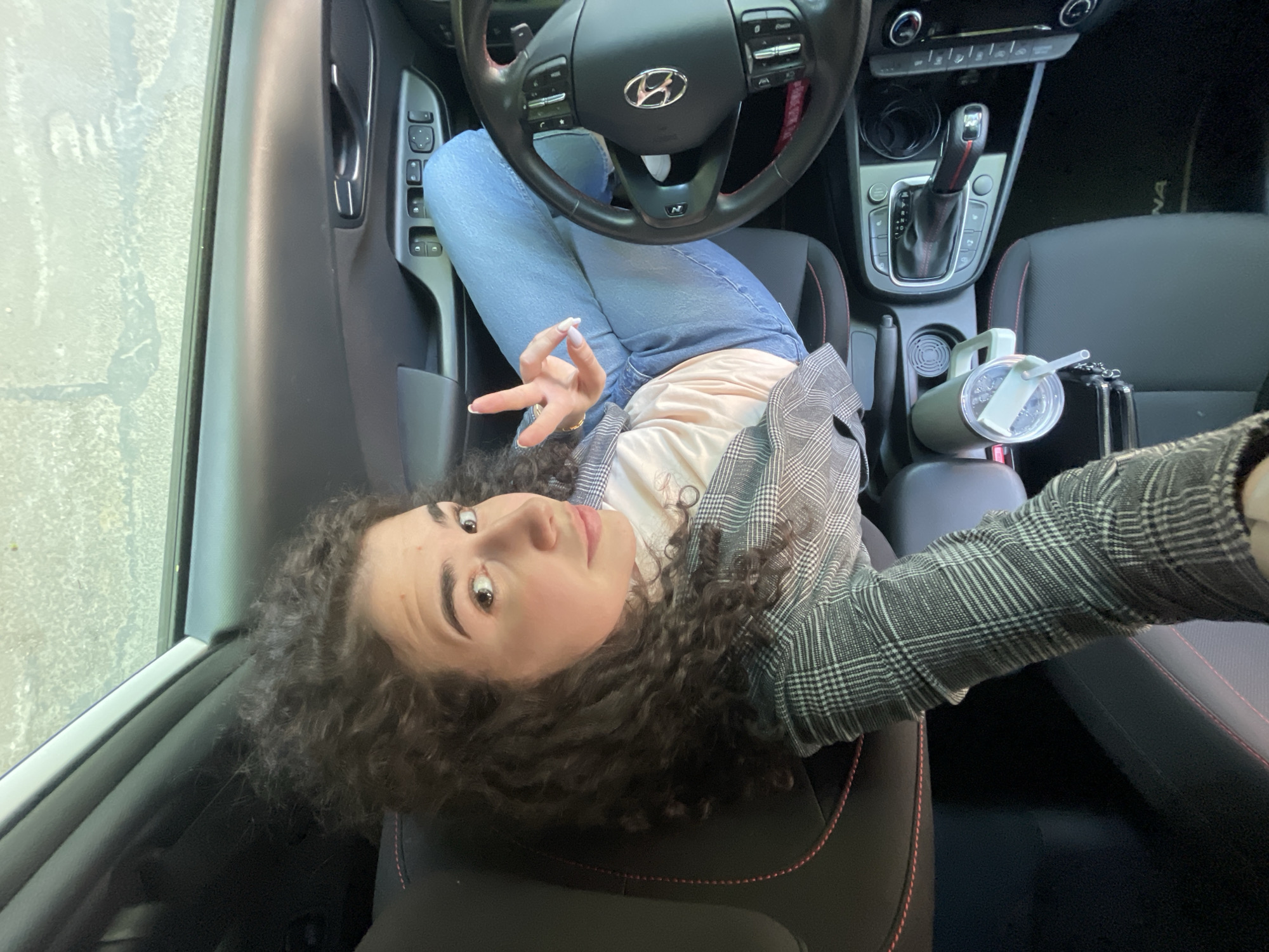 A woman is in the driver's seat of a car, angled back and looking at the camera. Her hair is curly, and she is wearing jeans, a pink top, and a plaid jacket. She is holding up two fingers in a peace sign. The car is a Hyundai, with an "N" logo on the steering wheel. There is a drink in the cup holder. The location is unknown, as there is no visual indicator within the provided scene to confirm it is in Sofia, Bulgaria.