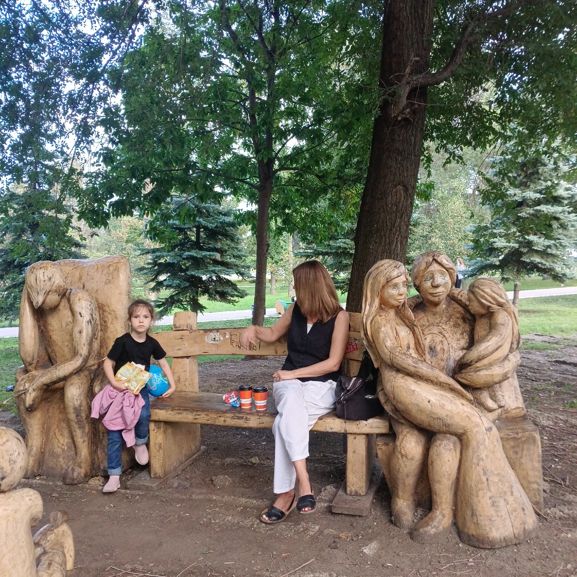 In Samara, Russia, a young girl in a black t-shirt and blue jeans sits on a wooden bench next to a large wooden sculpture of a grieving figure. She holds a bag of chips and a blue globe. To her right, a woman in a black vest and white pants also sits on the bench, looking to her right. Two disposable coffee cups and a small bag of candy are on the bench between them. To the right of the woman is a wooden sculpture depicting a family with two adults and a child. The scene takes place outdoors in a park-like setting with trees and grass.