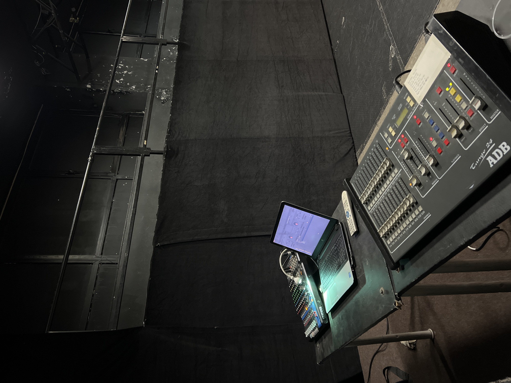 This media file captures a control station within a dark indoor environment, indicative of a theater or event venue control booth in Sofia, Bulgaria. The primary subject is a dark-grey control console, prominently labeled "Tango 24 ADB," positioned on a black surface. This console features an array of faders, illuminated buttons (red, yellow, green), and toggle switches, with discernible labels including "MASTER," "EFFECT," "PLAYBACK," "MANUAL," "GENERAL FUNCTIONS," and "RECORD."
Adjacent to the large console, a laptop is open, its screen displaying what appears to be control software. A smaller "YAMAHA" branded mixer, equipped with additional faders and buttons marked with channel numbers such as "6:18," "7:19," and "12:24," sits below the laptop. A white remote control is also visible near the laptop.
The background is dominated by dark, possibly fabric-covered, walls or stage structures, with some areas showing signs of wear and peeling paint on a metal framework. The overall lighting is dim, highlighting the glowing screens and indicator lights of the equipment, suggesting the setup is active for an event or in a preparatory phase. No people are visible in the scene.