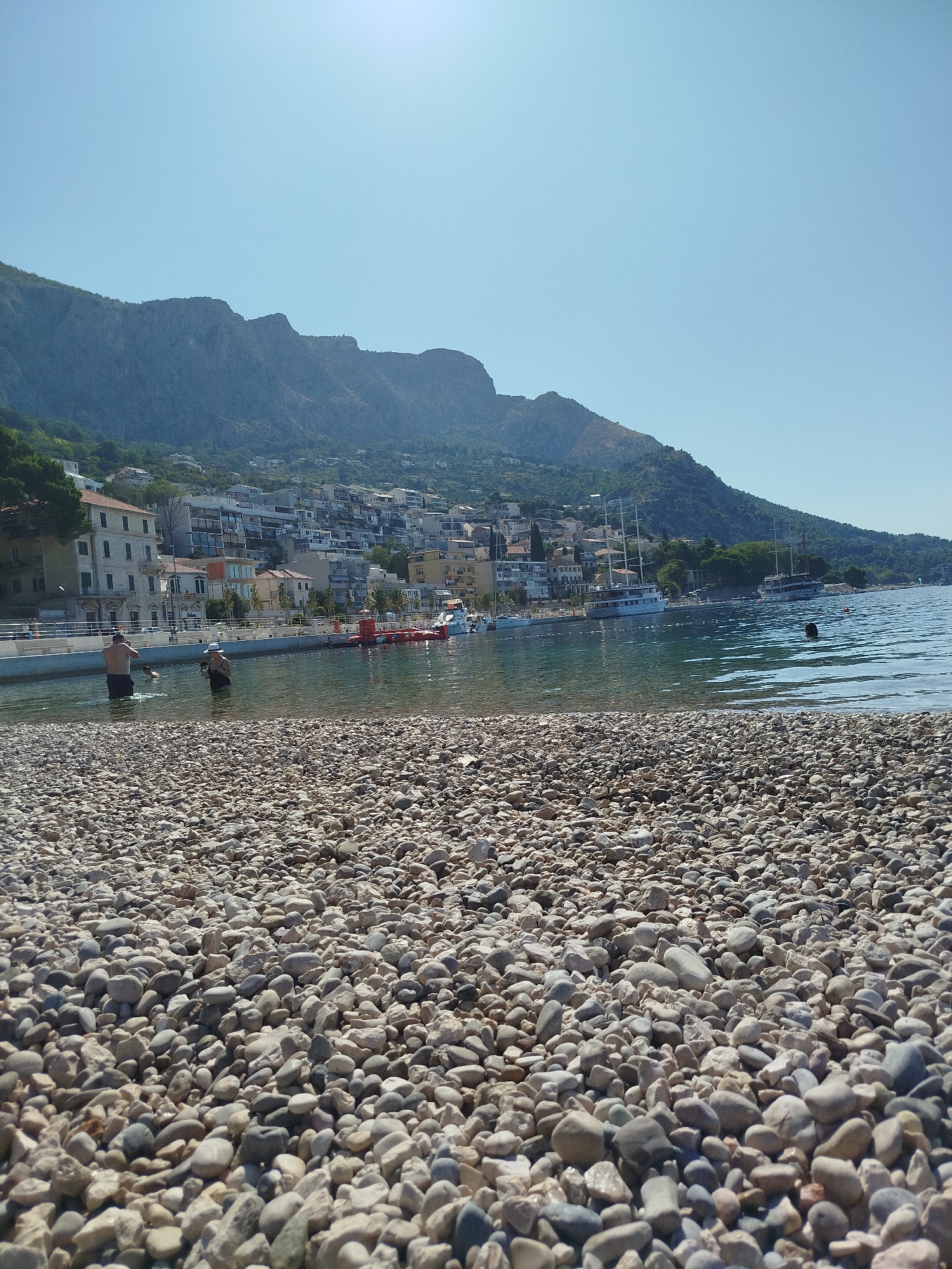 In Omiš, Croatia, this image captures a sun-drenched day at a pebble beach leading into the Adriatic Sea. In the foreground, a dense scattering of various sized, light-colored pebbles blankets the beach. A few people are visible wading in the clear, calm water. To the left, a man stands facing away from the camera, with his hands near his face as if shading his eyes or adjusting his hat. To his right, another person, possibly wearing a hat, stands in the water. In the mid-ground, the water extends to a promenade lined with buildings that ascend a lush, mountainous backdrop. Several boats are moored in the bay, including two large sailing vessels with white sails furled. The sky is a clear, bright blue.
