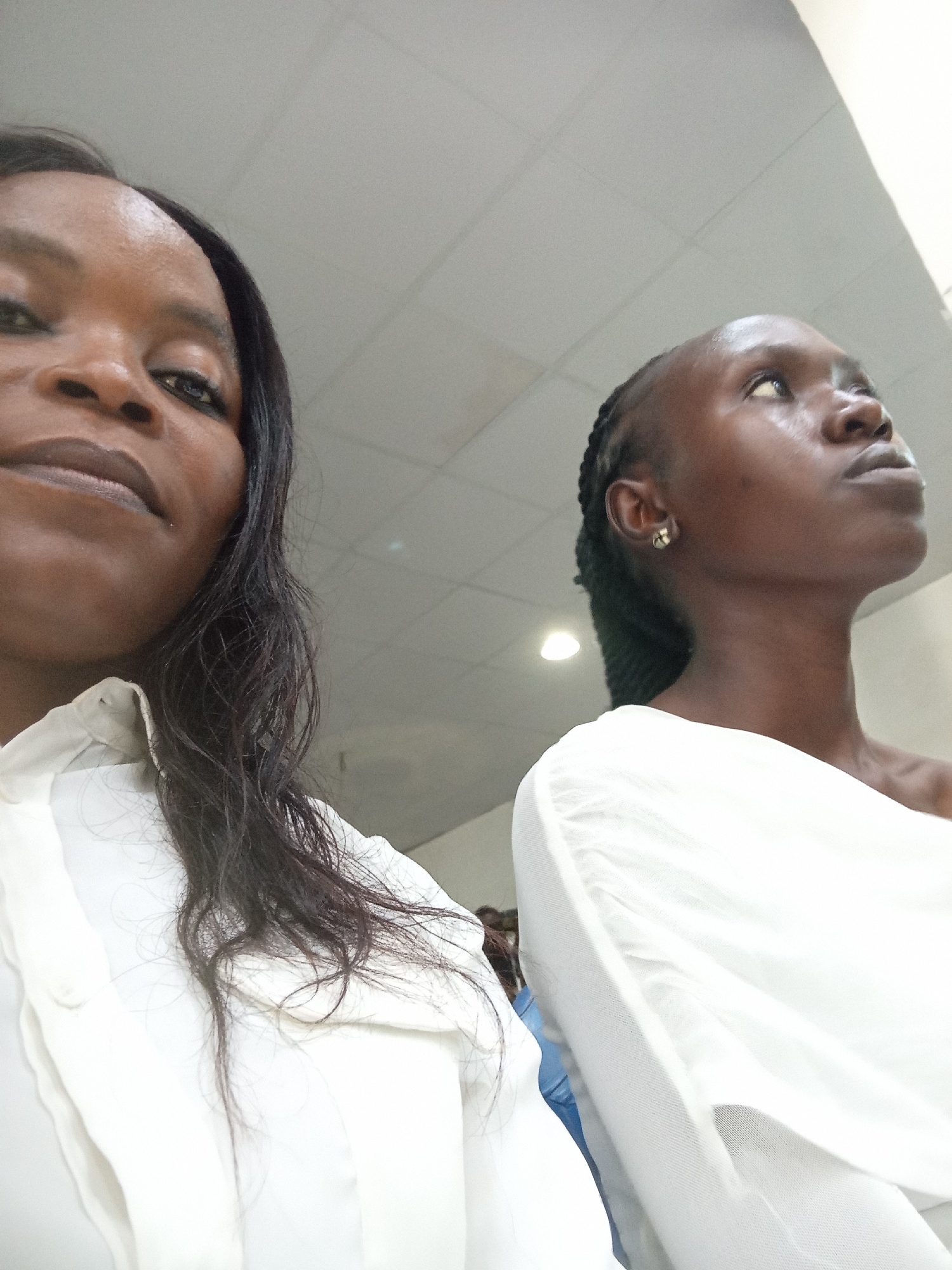 Two individuals are visible in a photograph taken from a low angle, looking upwards towards a white tiled ceiling. The person on the left is closer to the camera, her face and upper body in the foreground. She has dark skin, dark hair, and is wearing a white collared shirt. Her gaze is directed downwards and slightly to the left. The second person, on the right, is partially obscured by the first. This individual also has dark skin and appears to be looking upwards and to the right. They are wearing a white garment that drapes over their shoulder. The lighting suggests an indoor environment, possibly a public building. The scene is devoid of any specific Kason, Nigeria landmarks.
