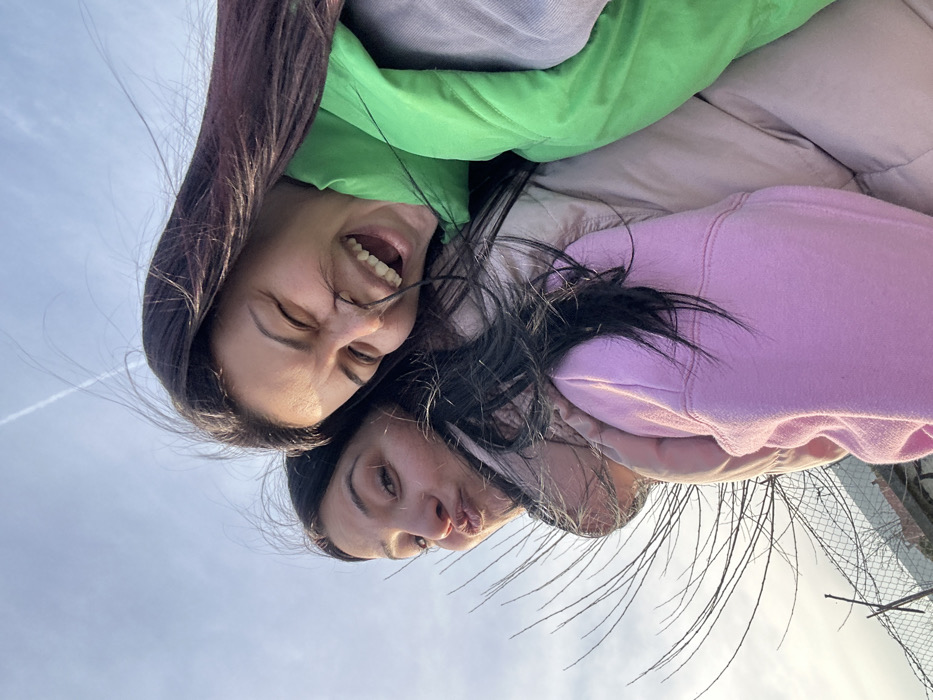 Two young adult females are depicted in a close-up, upward-angled photograph taken outdoors in Chubra, Bulgaria, under a mostly clear blue sky with a faint aircraft contrail. The subject on the right, possessing long dark hair, is shown in profile with a wide-open mouth, laughing, and exhibiting prominent teeth. She is wearing a bright green padded vest over a lighter-colored top. The subject on the left, also with long dark hair, faces more directly towards the camera, displaying a playful pouting expression with pursed lips. She is dressed in a light pink hooded sweatshirt over a light beige or pink padded jacket. Both individuals' hair appears windswept. They are positioned in close physical proximity, suggesting a shared moment or embrace. In the lower-left background, bare tree branches and a chain-link fence topped with barbed wire are partially visible, indicating an outdoor setting adjacent to an undeveloped or contained area.