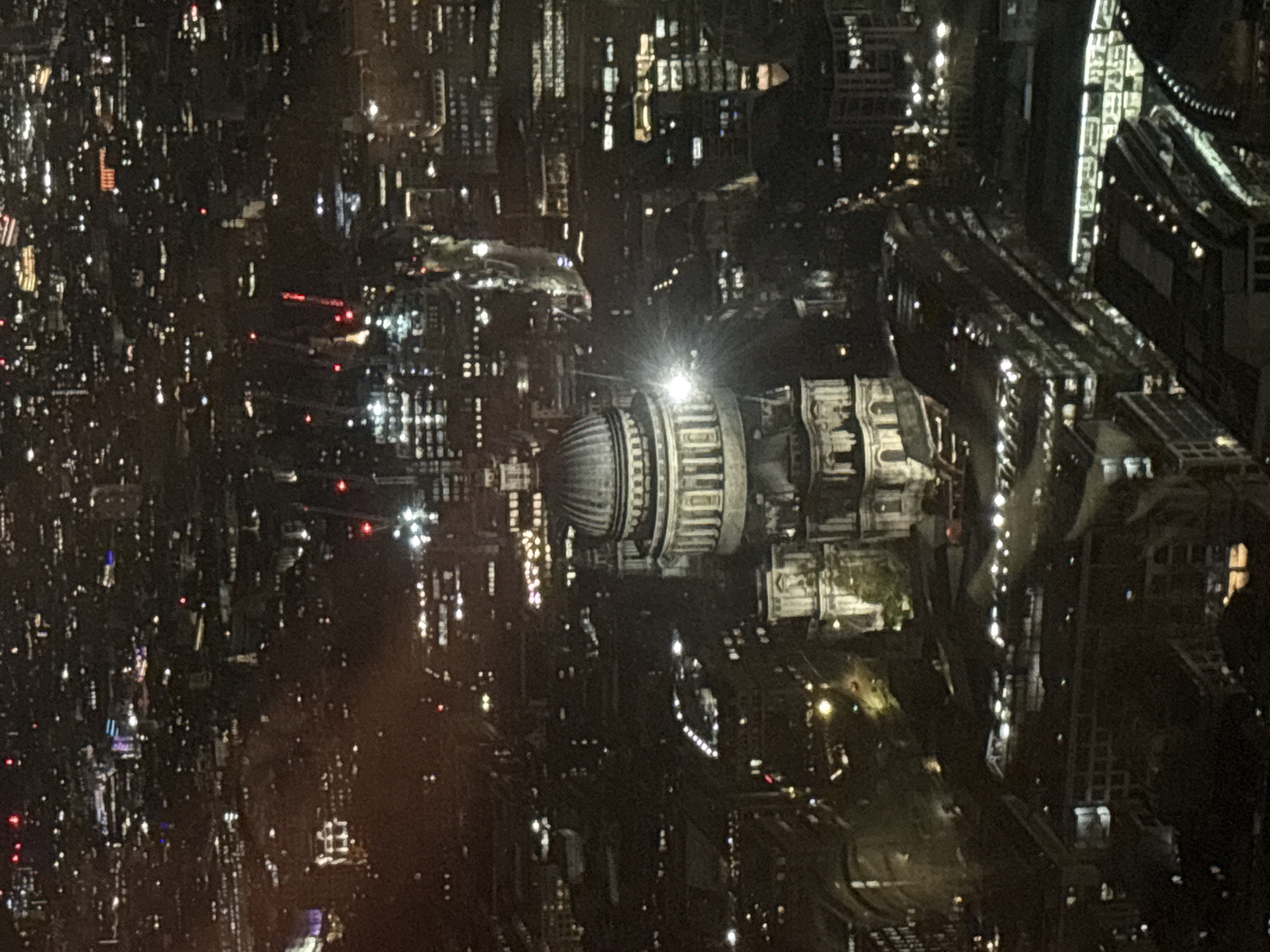 An aerial night shot of London, United Kingdom features the dome and structure of St. Paul's Cathedral illuminated in the center. Reflections of city lights twinkle across the surrounding urban landscape. The image captures the intricate web of streets and buildings within the city.