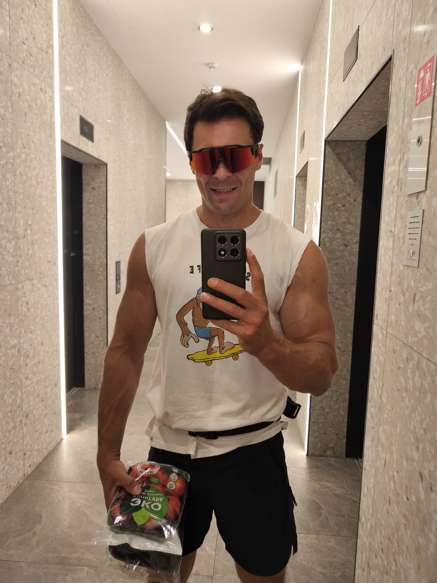 A man wearing a white sleeveless t-shirt, black shorts, and red sunglasses is standing in a well-lit hallway with elevators. He is holding a package of plums in his left hand and a smartphone in his right hand, taking a selfie. The hallway has beige speckled walls and marble floors, with vertical lighting strips. The elevators are dark-colored. The man has muscular arms and a smiling expression.