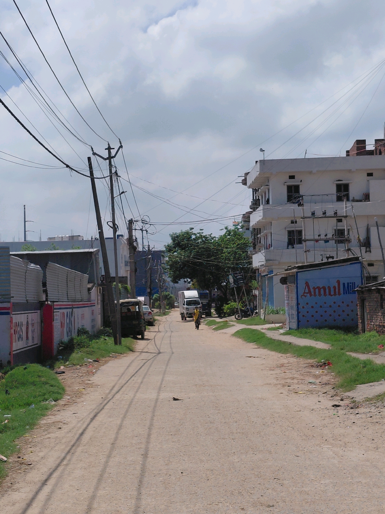 The image depicts a street scene in Patna, India. The street is unpaved and dusty, with tire tracks visible on the surface. Overhead, a complex network of electrical wires crisscrosses the sky, supported by utility poles. On the left side of the street, a corrugated metal structure with signage that reads "HP GAS" is visible. Further down the street, a small, open-air vehicle, possibly a tuk-tuk, is parked.
On the right side of the street, a multi-story white building with multiple windows dominates the view. Below this building, a smaller structure with a blue facade displays the "Amul Milk" logo. A few trees are interspersed along the street.
In the distance, a few individuals are present. One person, wearing a yellow garment, is walking down the street. Other figures are less distinct, but suggest ongoing activity. The sky is overcast with clouds, indicating a possibly humid or monsoon season day. The overall impression is of a lived-in urban environment with active infrastructure and daily life.