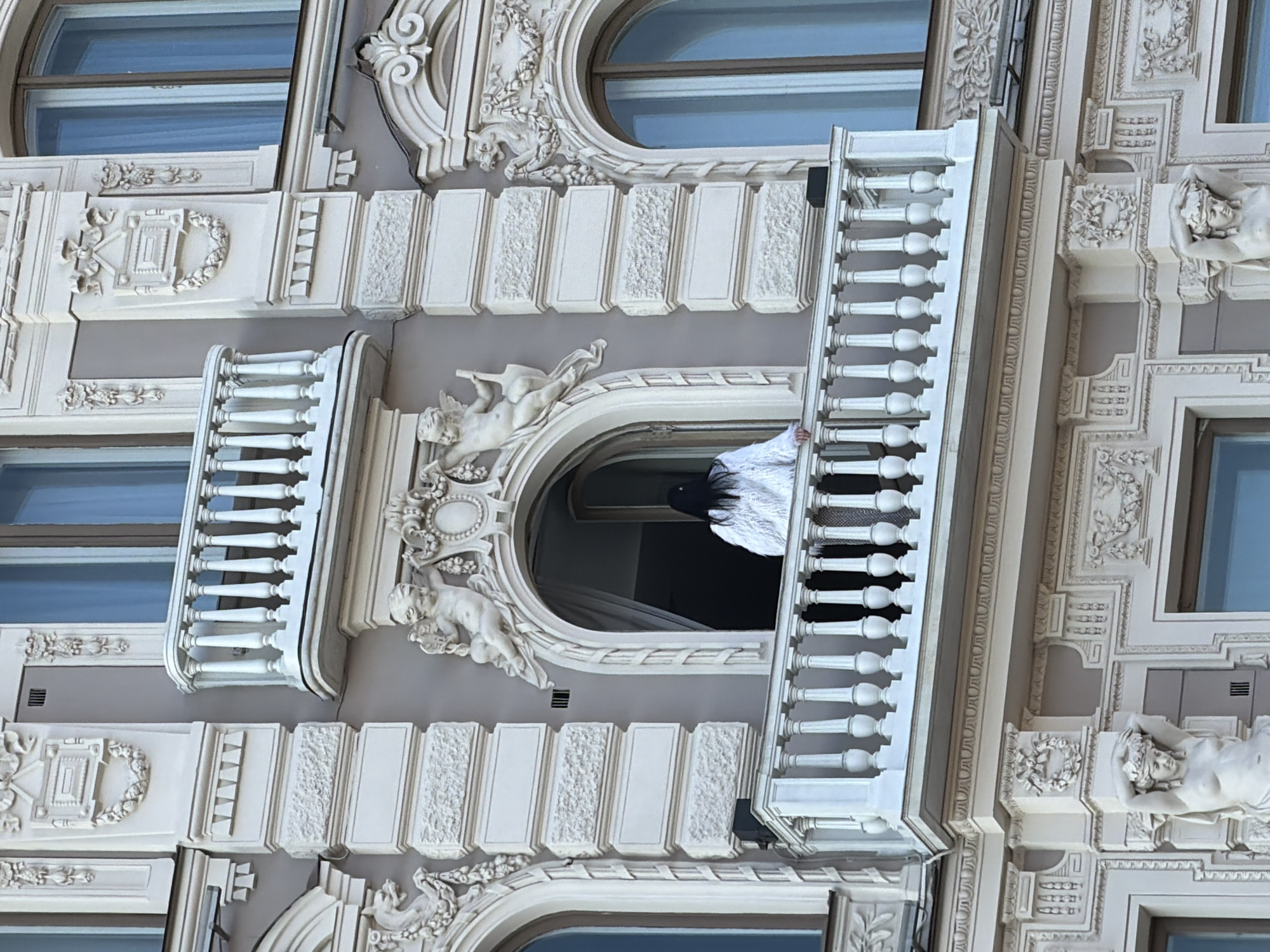 The image shows a building facade in Saint Petersburg, Russia, with ornate architectural details including sculptures of cherubs and reclining figures, relief patterns, arched windows, and balustraded balconies. A person wearing a white garment and a black head covering is visible on one of the balconies.