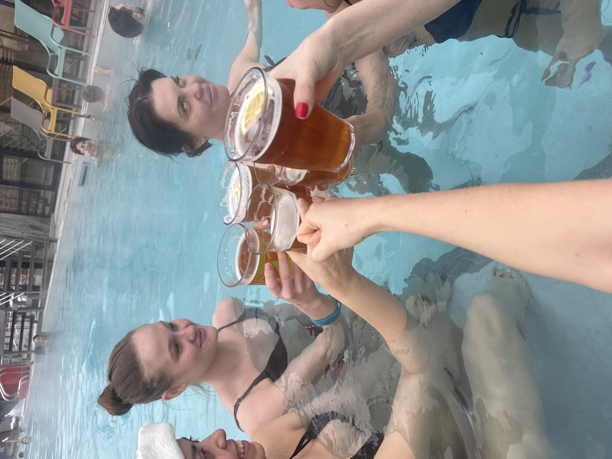 The image captures a group of women in a pool, toasting with glasses of amber liquid, possibly beer, some with lemon slices. The pool appears to be indoors, based on the structure and seating visible on the left side. Steam rises from the water, indicating it is heated.
The location cannot be definitively determined as Moscow, Russia from the information provided.