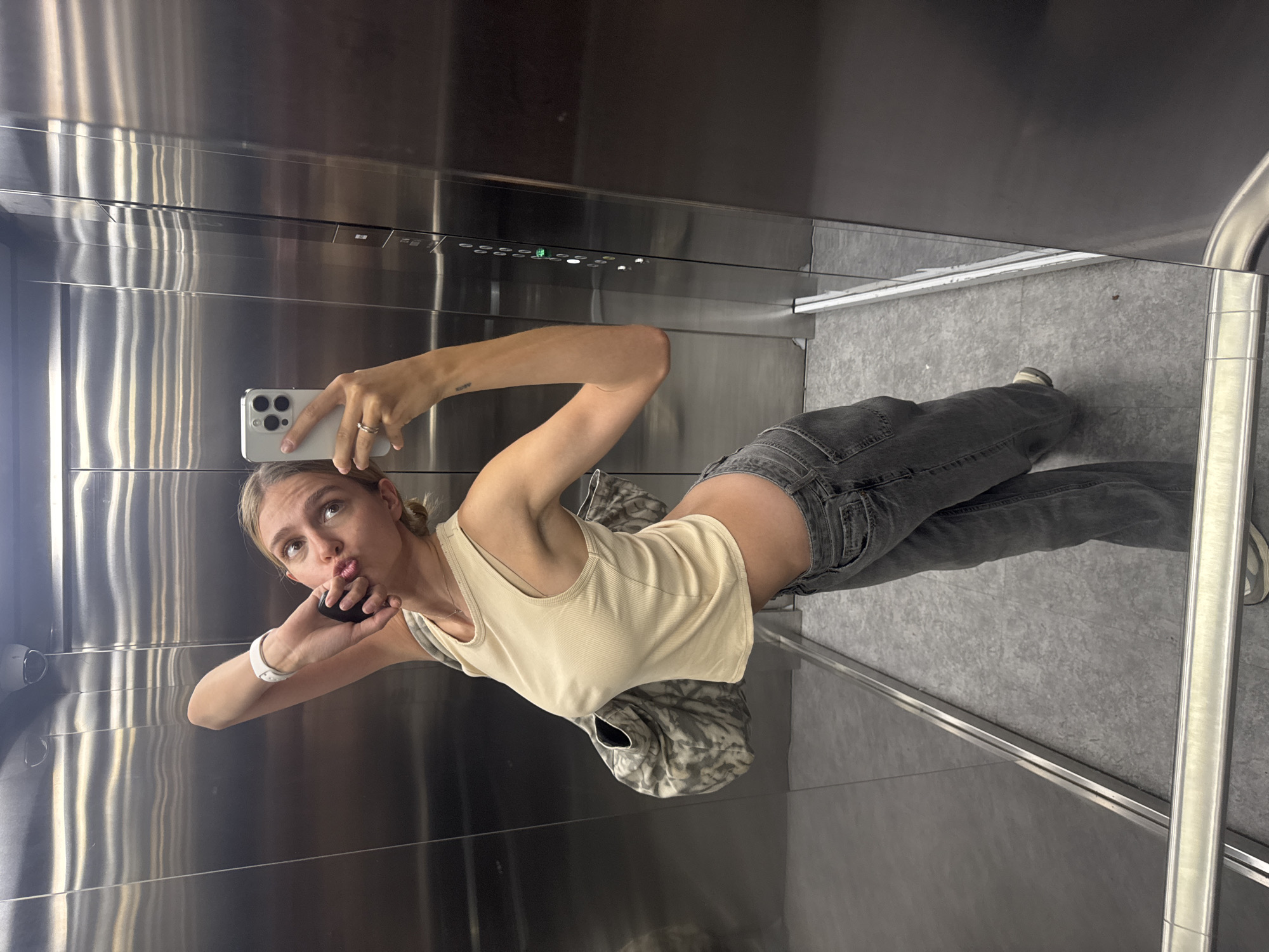 A young woman takes a selfie in an elevator with reflective metal walls and a grey tile floor. She wears a light tan tank top, dark grey jeans, and a white watch. She's holding a white smartphone in her right hand. A grey backpack rests on the floor near her. The location appears to be an interior space, specifically an elevator, in a city of unknown location.