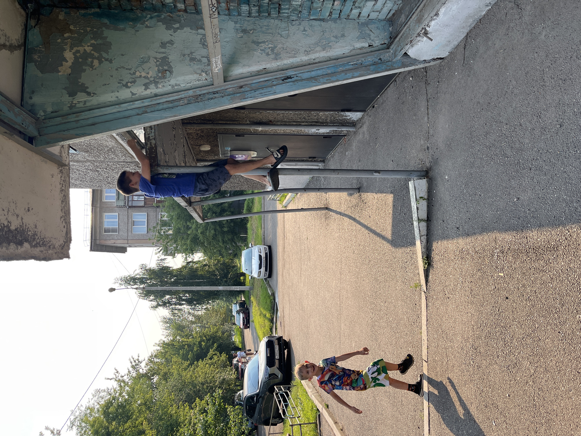 In Krasnoyarsk, Russia, on a sunlit day, two young boys are visible. One boy, wearing a blue t-shirt and dark shorts, is climbing on the metal supports of an elevated, weathered, turquoise-painted awning attached to a building with a rough concrete facade. He is barefoot. The other boy, wearing a colorful t-shirt with cartoon characters and camouflage-patterned shorts, is running on an asphalt pathway. He is wearing dark sneakers and socks. Behind them, several cars are parked in an outdoor area with green grass and trees. The building itself appears to be a multi-story apartment complex with visible windows. The overall scene depicts a common urban residential environment.
