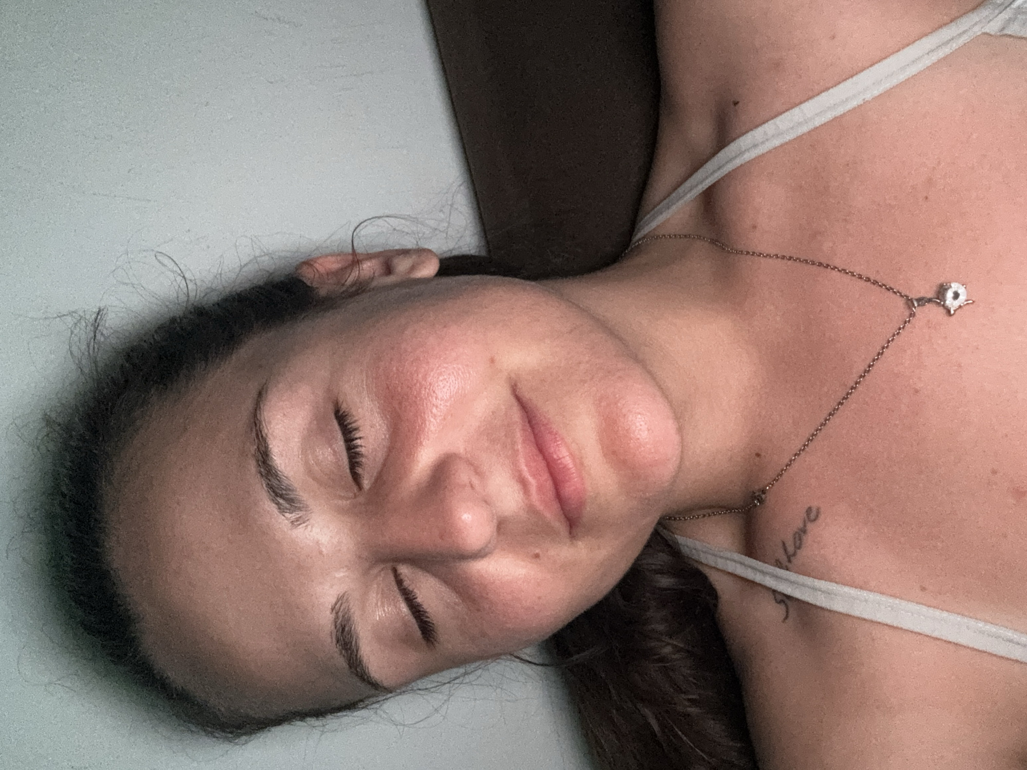 The image is a close-up, overhead shot of a young woman with her eyes closed, resting her head against a dark brown surface, possibly a couch or pillow. She is facing slightly to the left, with her hair pulled back. Her face is illuminated by a soft light, highlighting a blush or redness on her cheeks. She is wearing a light-colored tank top, and a silver necklace with a small, light-colored pendant is visible around her neck. There is a small tattoo on her collarbone that appears to read "Sh Love." The background is a plain, light-colored wall. There is no discernible indication of the city of Plovdiv, Bulgaria, or any other specific geographic location.