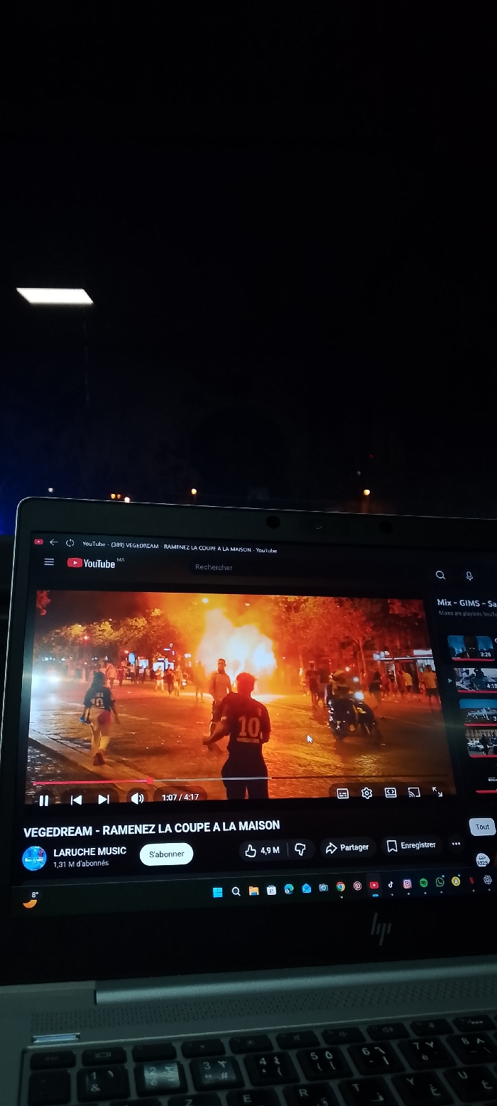 A laptop displays a YouTube video titled "VEGEDREAM - RAMENEZ LA COUPE A LA MAISON." The video shows a street scene with a burning object, possibly a vehicle, with people nearby. Two figures wearing football jerseys are prominent in the foreground. The YouTube interface is visible, including playback controls, channel information ("LARUCHE MUSIC"), and various icons for liking, sharing, and saving the video. The laptop's keyboard is also in view.