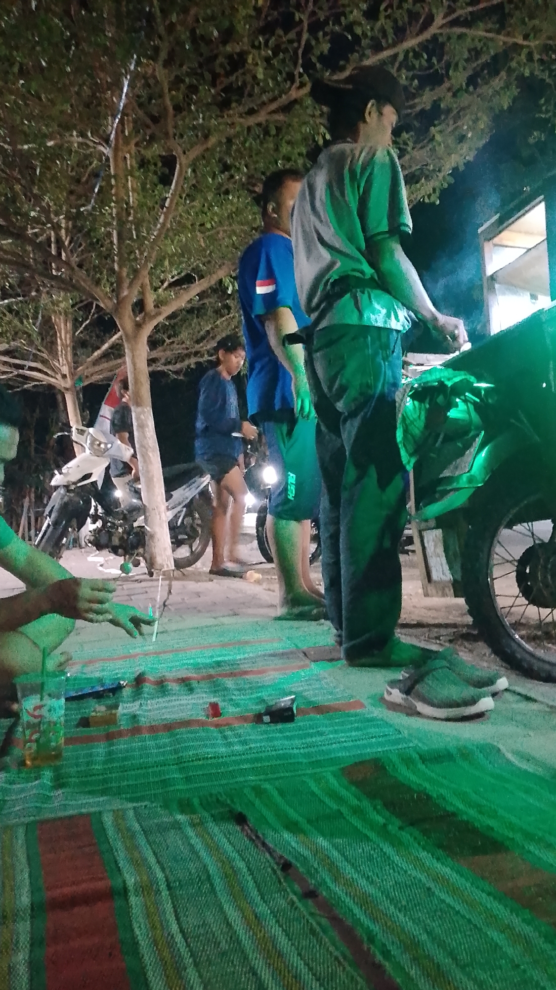 The image depicts an outdoor scene at night, likely in a residential or public area in Karanglangu, Indonesia. Several individuals are present, with varying distances and levels of detail. In the foreground, a person is partially visible, sitting on a woven mat. The mat, predominantly green with red and yellow stripes, covers the ground. A plastic cup with a straw and a dark liquid is placed on the mat, alongside a metallic object that may be a lighter or a pen.
Further into the image, three men are standing. One man, wearing a blue t-shirt with a white and red stripe and dark shorts, is positioned centrally. To his right, another man is seen from the side, wearing a grey t-shirt and dark trousers. His face is obscured. Behind them, partially visible, is a motorcycle, and another is parked further back. The environment includes trees with foliage, casting shadows and creating a natural canopy. The lighting is dim, with some areas illuminated by artificial light, casting a greenish hue on parts of the scene, particularly the motorcycle. The overall impression is of a casual gathering or break during nighttime.