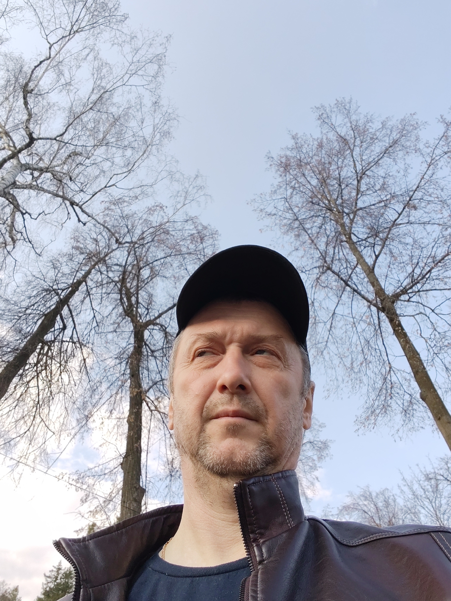 A medium-shot selfie captured outdoors shows a middle-aged man wearing a black baseball cap and a brown leather jacket over a dark blue t-shirt. He has short graying hair and a trimmed beard. The man's gaze is directed towards the upper right. Behind him, bare trees stretch upwards against a pale blue sky, suggesting it is either late autumn or early spring. The trees appear to be located in a park or wooded area, and given the provided location, it would likely be a city park in Sarapul, Russia. More specifically, based on tree type and open sky, it is likely the park area near the Kama river.
