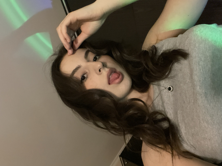 A young woman with long, wavy dark hair poses provocatively. She wears a gray top and a necklace with a circular pendant. Her hand is near her forehead, and she sticks out her tongue. The background features a white wall with green light streaks. The overall image has a casual, selfie-like quality.