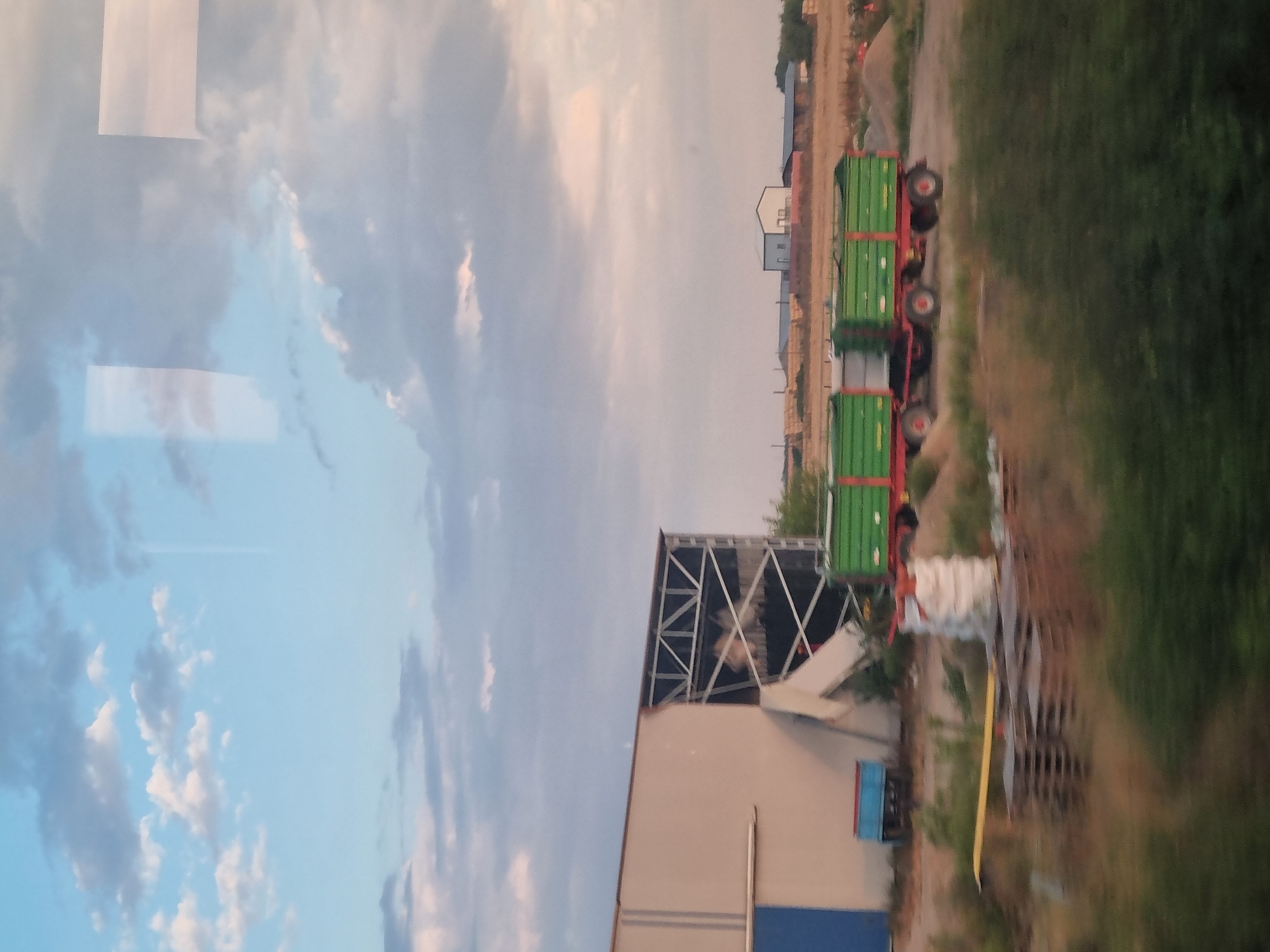 In Ciumeghiu, Romania, an industrial area is depicted under a cloudy sky. A large, open-sided building with exposed metal beams is visible on the left. In front of it, a blue trailer is parked. A red tractor-trailer with two green wagons sits on a dirt road in the middle ground. Piles of dirt and hay bales are scattered around the area, and a large white bag is near some wooden pallets. In the background, several industrial buildings and a distant town are visible. The foreground is filled with blurred green foliage.