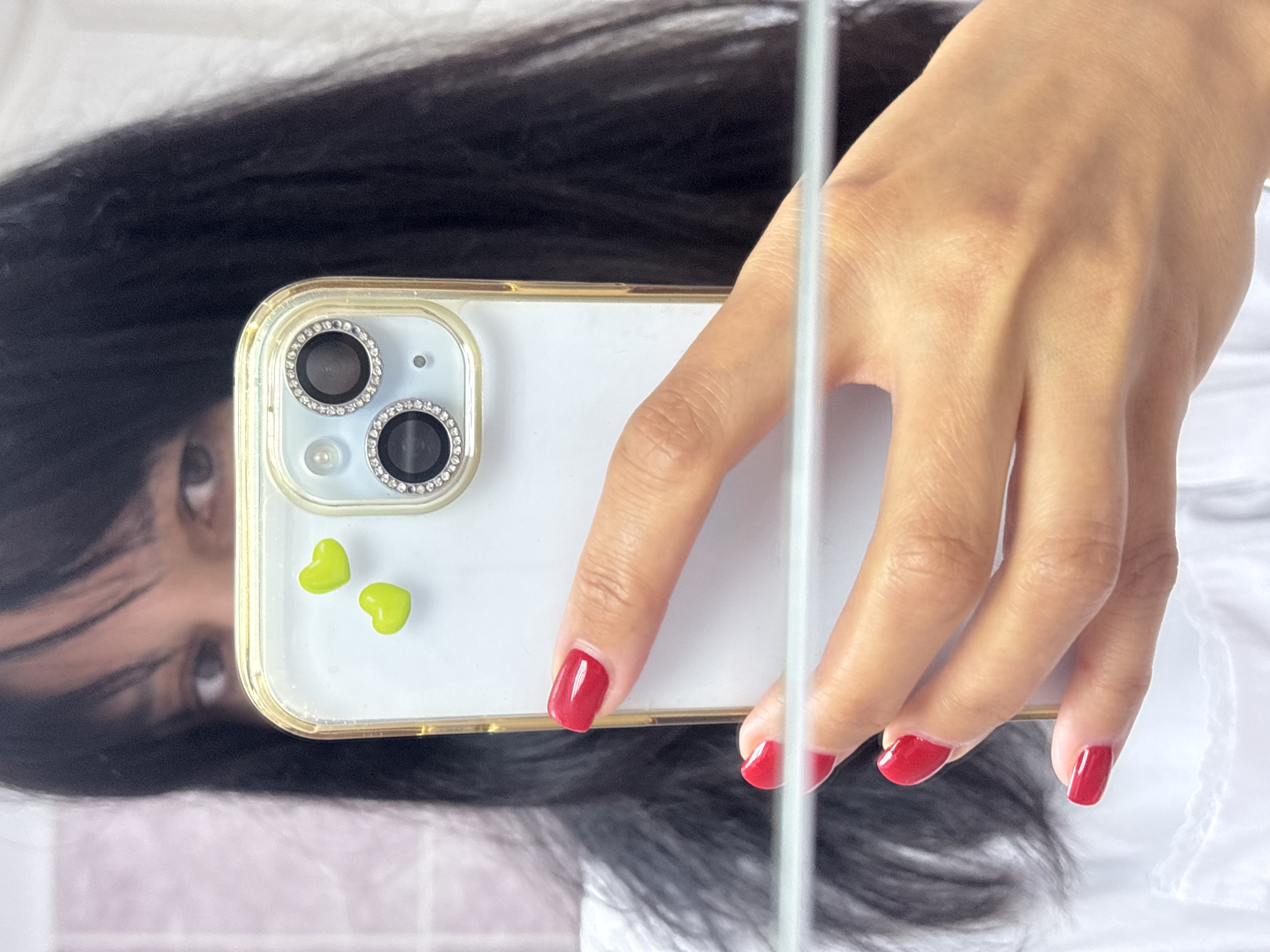 A selfie taken in a mirror shows a person holding a white phone with two green heart-shaped decorations and a jeweled camera surround. The person has dark hair, visible eyes, and is wearing a white shirt. The person's hand is visible, with red nail polish. The scene location is impossible to determine from the image.