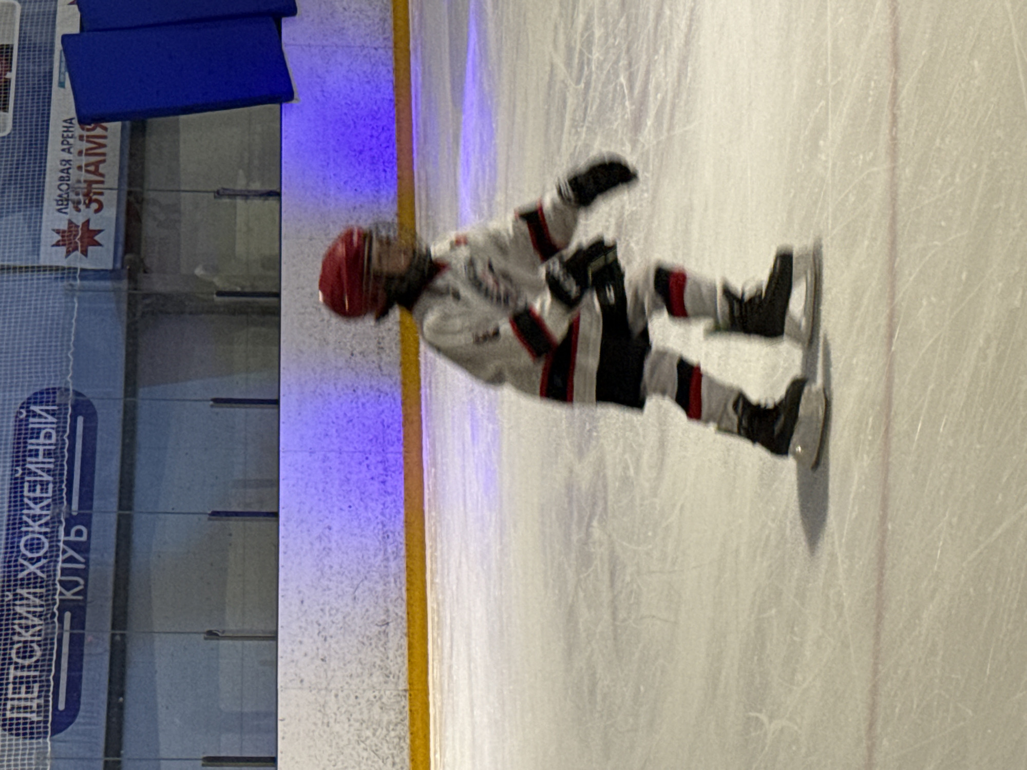 The image depicts a young person wearing hockey gear, including a red helmet and a white jersey with red and black trim, on an ice rink. The person is in motion, suggesting they are skating or playing hockey. The background shows an ice arena with blue padding and signage. The signage in the background is in Russian and translates to "Children's Hockey Club" and "Ice Arena Znamya". This indicates the location is likely the Ice Arena Znamya in Votkinsk, Russia, which is a known facility for youth hockey. The lighting in the arena appears to be artificial, with some purple-hued lights visible. The ice surface shows visible skate marks.