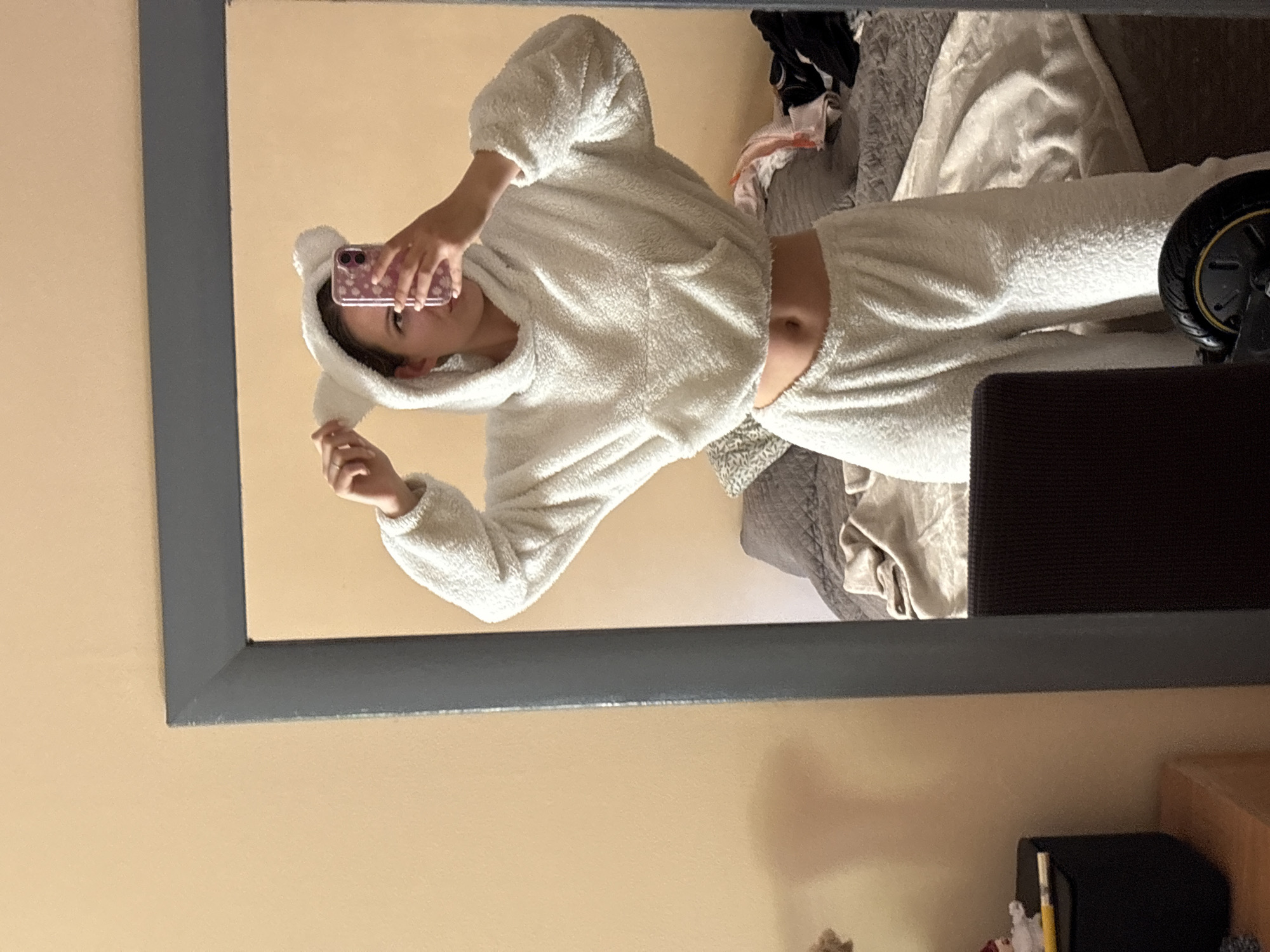 An individual takes a mirror selfie in what appears to be a bedroom. The person, likely female, wears a matching two-piece loungewear set made of white, fluffy material. The top is a cropped hoodie with small ear-like attachments on the hood, exposing the individual's midriff. The bottom piece consists of matching sweatpants. They hold a smartphone with a pink case, patterned with what looks like small hearts or flowers, which partially obscures their face, leaving only part of their eyes visible.
The large rectangular mirror has a dark grey frame. The room's wall reflected in the mirror is a uniform, warm beige or light yellow color. In the background of the reflection, behind the person, a bed is visible with rumpled bedding in shades of grey and white, along with some piled clothing. To the right of the person's reflection, a dark brown piece of furniture, possibly a headboard or cabinet, is seen, along with a black object featuring a yellow rim, potentially a wheel from a scooter or similar item. In the lower right corner of the reflected room, a light wooden surface is visible, alongside a small black object and what appears to be a yellow pencil or similar writing implement. The setting suggests a casual, private indoor space. The location context provided is Bečići, Montenegro, though there are no specific visual cues within the image to indicate this. No discernible text is present in the image.