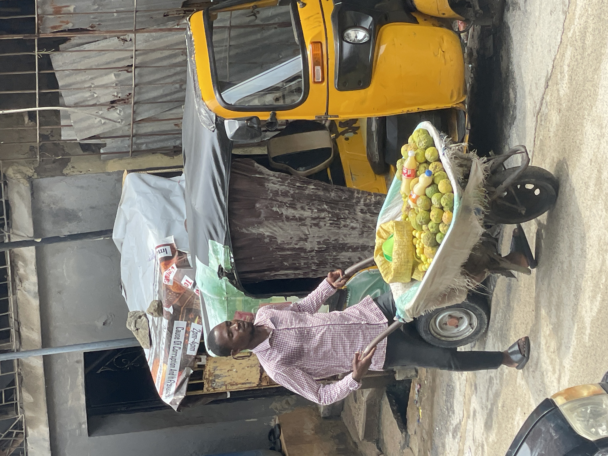The image depicts a man in Lagos, Nigeria, positioned next to a yellow auto-rickshaw (a common form of transportation in Lagos). The man, dressed in a checkered shirt and dark pants, stands on a stepped curb holding a two-handled cart. The cart is filled with green-colored fruits, some bottles, and a yellow cloth. An advertisement poster with visible words "Cause Of Corruption And Its Prevent" is attached on the cart. The surrounding area appears to be a street in Lagos, with corrugated metal walls and other vehicles visible.