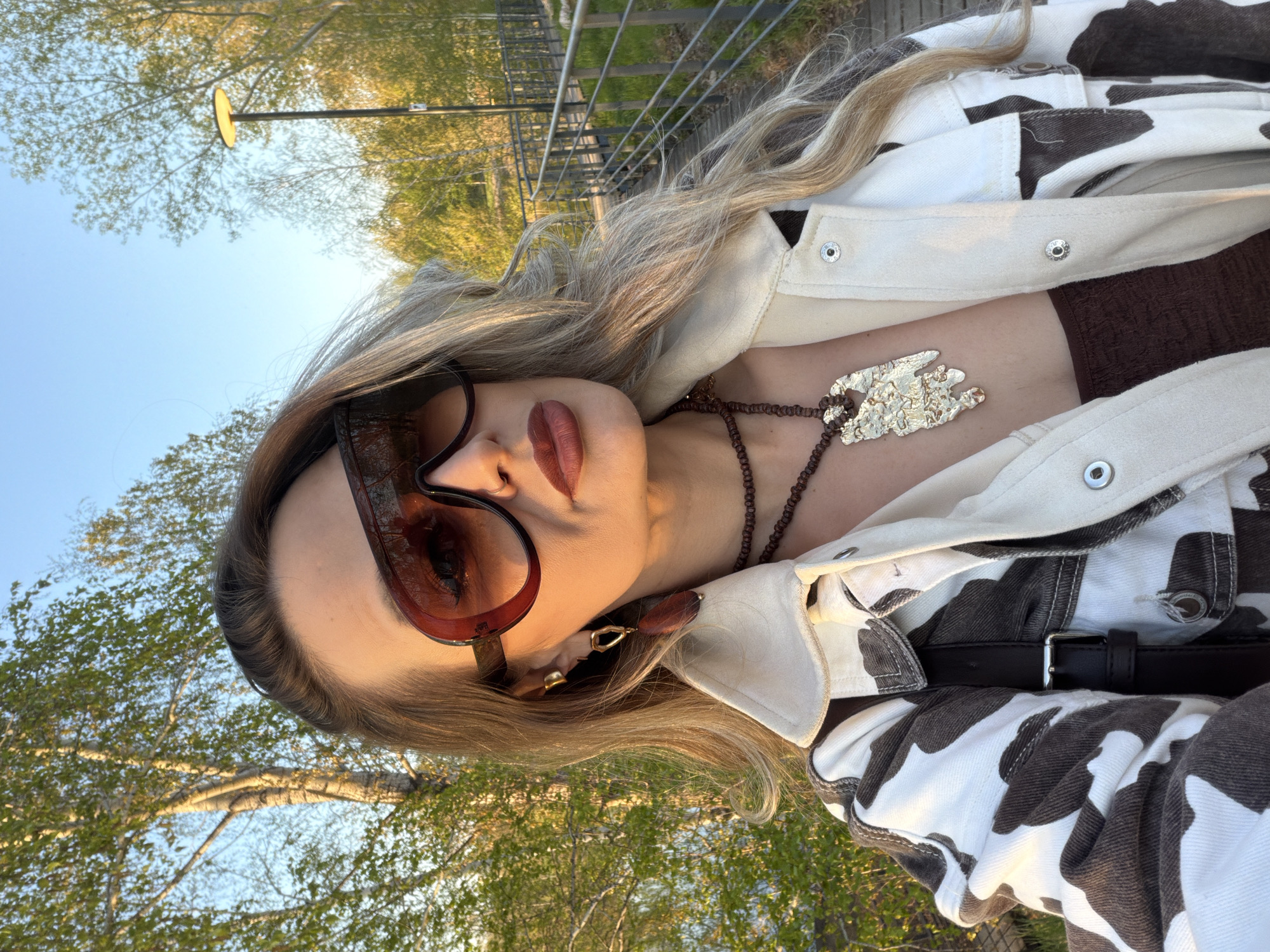 A blonde-haired female is depicted in a close-up outdoor shot, likely a selfie, against a backdrop of trees and a clear sky. She is wearing large, tinted sunglasses, a brown top, a brown beaded necklace with a silver pendant, and a cow-print jacket, and a black belt is visible on her waist. The location appears to be Krasnoyarsk, Russia, based on visual cues like the architectural style of railings and lamp posts that can be found in the city. The lighting suggests it is either early morning or late afternoon. She appears to be standing near a path or bridge with railings.