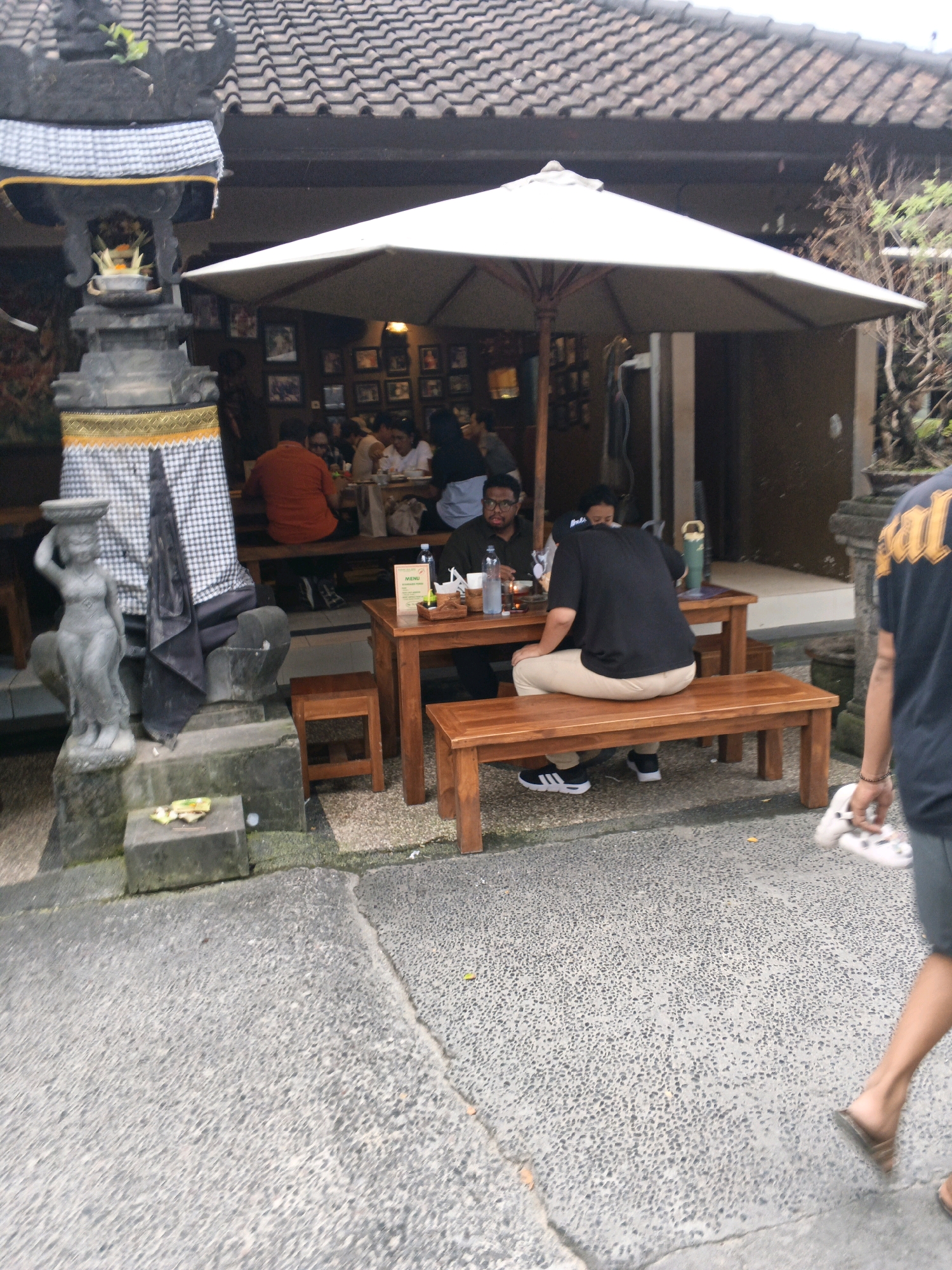 The image shows an outdoor seating area of a restaurant in Denpasar, Indonesia. A thatched roof covers the building in the background. In the foreground, people are seated at wooden tables and benches under a large white umbrella. A statue of a woman carrying a bowl on her head stands to the left of the seating area, next to a stone structure with a patterned cloth draped over it. The ground is paved with asphalt. A person is walking away from the camera on the right side of the image, carrying a pair of sandals. The overall scene depicts a casual dining environment with traditional Balinese architectural elements.