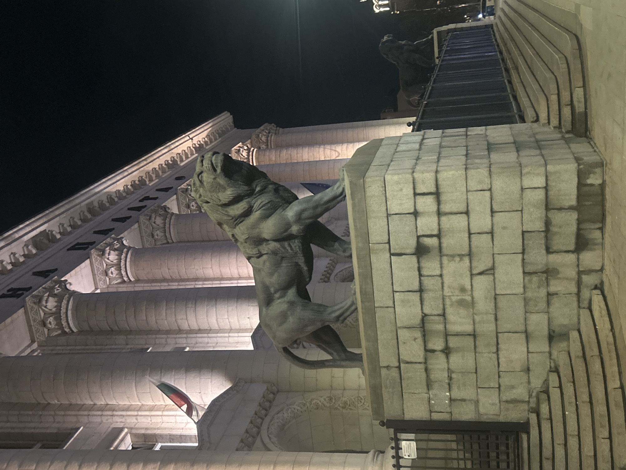 The image depicts the Palace of Justice in Sofia, Bulgaria, at night. A large stone lion statue stands prominently in the foreground, mounted on a stone pedestal. The classical architectural features of the building are visible, including columns, ornate carvings, and a portion of the Bulgarian flag. In the background, steps leading upwards are observed. The lighting highlights the details of the architecture and statue.