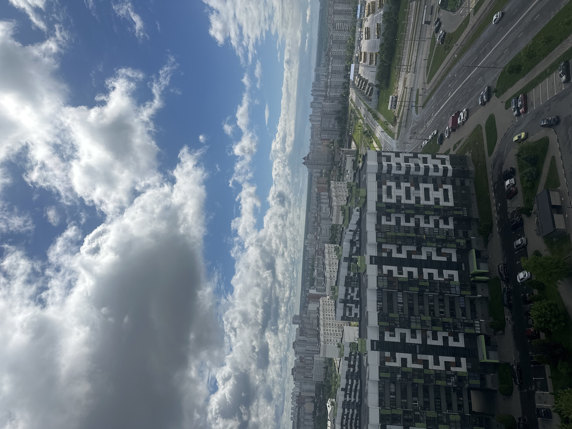 The image depicts a high-angle view of a cityscape under a partly cloudy sky. In the foreground, there's a tall building with a black and white patterned facade. Adjacent to the building is a road with multiple lanes, cars parked along the side, and trees lining the street. In the background, more buildings stretch into the distance, fading slightly due to atmospheric perspective. The sky is blue with scattered white and grey clouds. The image appears to be taken in or around the city of Бараўлянскі сельскі Савет, Belarus.