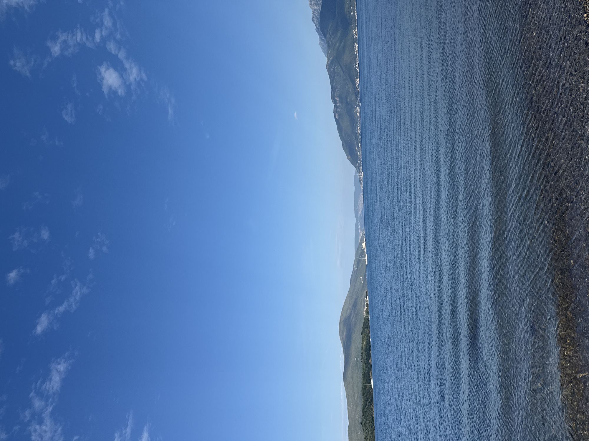 This image depicts a tranquil coastal scene in Mrčevac, Montenegro, under a clear blue sky dotted with scattered white clouds. The foreground features the shallow, clear waters of the Adriatic Sea lapping against a pebble-strewn shore. Gentle ripples on the water's surface indicate a calm day. In the mid-ground, a wide expanse of blue sea stretches towards rolling green hills and distant mountains that frame the horizon. Several sailboats and yachts are visible on the water, suggesting a popular location for sailing. The landscape is characterized by lush vegetation on the hillsides and scattered buildings nestled along the coast. The overall impression is one of natural beauty and peacefulness.