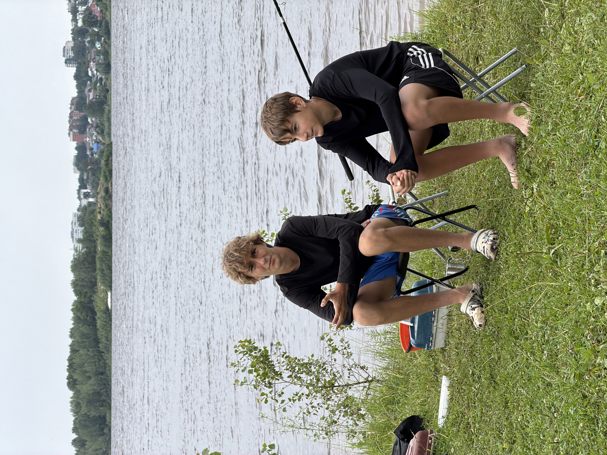 Two young males are sitting on folding chairs on a grassy bank overlooking a wide river. Both are wearing long-sleeved black shirts. The male on the left is wearing blue shorts and patterned clogs. The male on the right is wearing black shorts with white stripes and is barefoot. There is a fishing rod extending from the right of the frame, partially visible. In the background, across the river, is a treeline and a settlement with buildings. The sky is overcast. The location is identified as the city of Noviy, Russia.