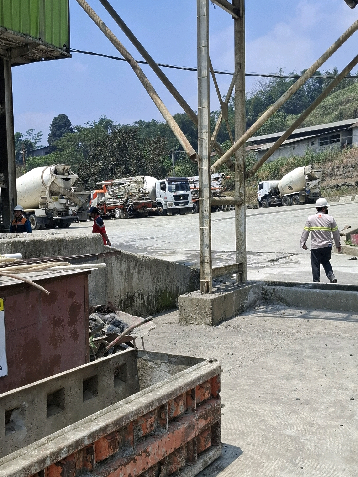 The image depicts a concrete mixing facility in Cijangkar, Indonesia. In the foreground, a man wearing a white hard hat and a reflective vest is walking away from the camera on a concrete surface. Behind him, a cement mixer truck is parked. Further in the background, several other cement mixer trucks are visible, some of which are stationary. The facility appears to be situated on a hillside, with vegetation and buildings visible in the distance. The sky is clear and blue. There are construction materials and equipment scattered around the site.