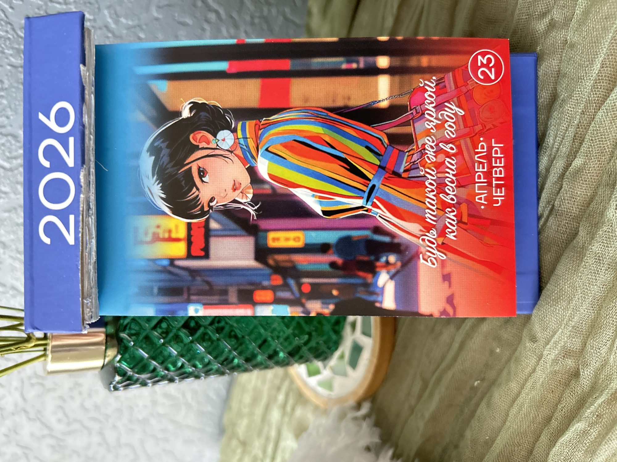 A 2026 calendar lies on a textured surface. The spine of the calendar is blue and displays the year "2026" vertically in white. The calendar is open, revealing a brightly colored page featuring an anime-style illustration of a young woman in a striped dress. The background of the illustration depicts a cityscape. The page is marked with "23" in a red circle in the upper right corner, and text in Cyrillic script, including "АПРЕЛЬ. ЧЕТВЕРГ", is visible. A green decorative object is visible beneath the calendar. The surface is textured. The location of the scene is Saint Petersburg, Russia.