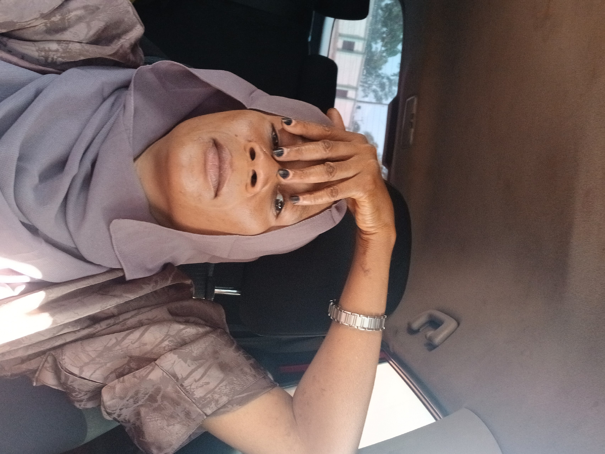 A woman wearing a light purple headscarf and a patterned brown top is seated in a car, partially covering her face with her hand. She is wearing black nail polish and a silver bracelet. The car interior has a gray headliner and black seats. Outside the car window, buildings and trees are visible, suggesting an urban setting. The image might have been taken in Rigachikun, Nigeria, but this cannot be verified without further information.