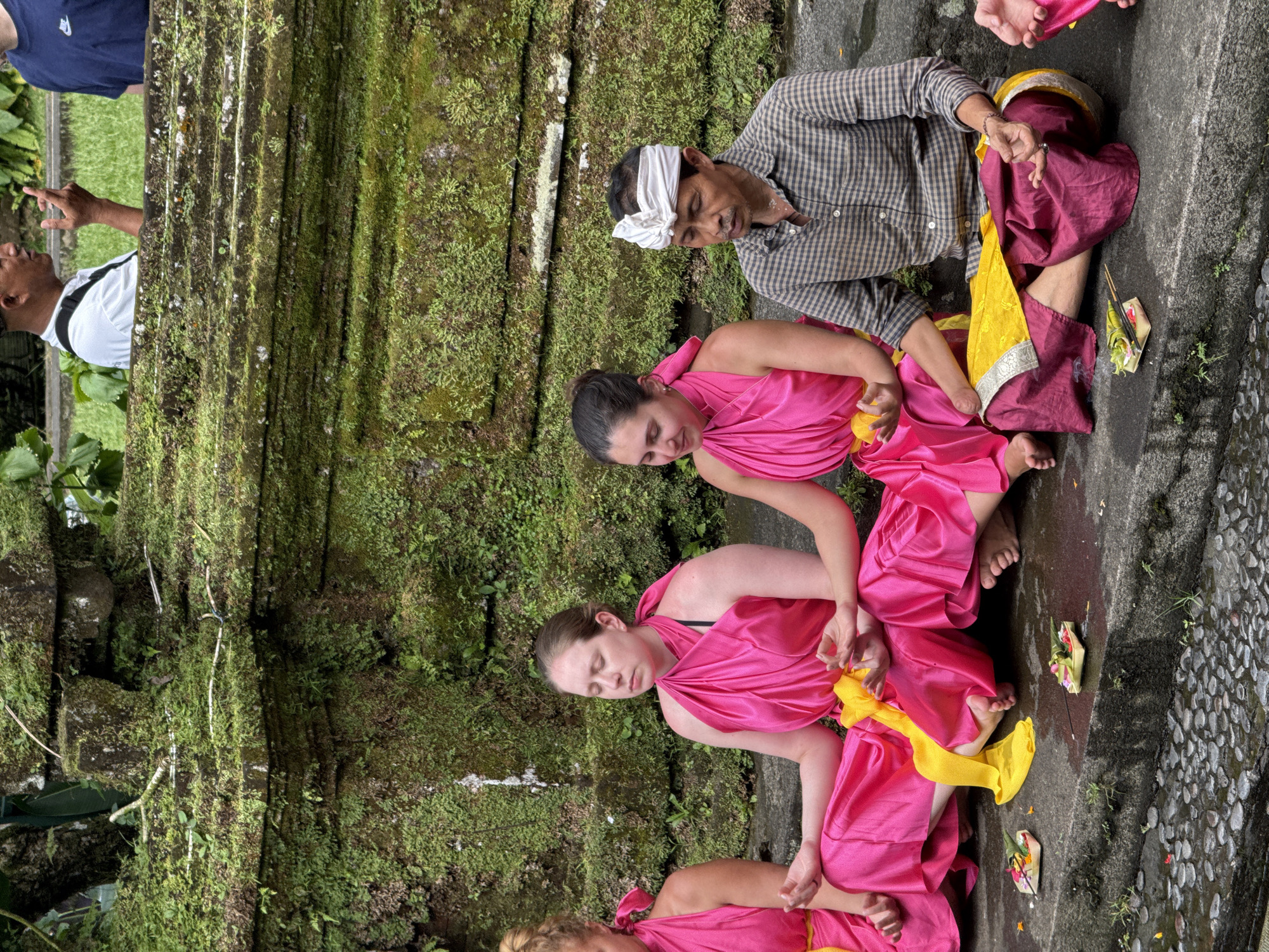 A gathering of people is captured in this outdoor scene, likely located in Tegallalang, Indonesia. Three individuals, two women and a man, are positioned close together on a stone surface. All three are in seated postures and seem to be engaged in an activity or ritual. The women are draped in bright pink fabric, with a yellow cloth incorporated into their attire. The man, situated adjacent to them, is dressed in a checkered shirt and has a white cloth wrapped around his head. Nearby, there are what appear to be small, intricate offerings, possibly related to the activity they are engaged in. The backdrop consists of a moss-covered stone wall, which hints at the site's age and the humid climate. Additional individuals are partially visible on the left of the frame, suggesting a larger group or onlookers. The overall impression is one of a cultural or religious activity taking place in a historic or natural site.