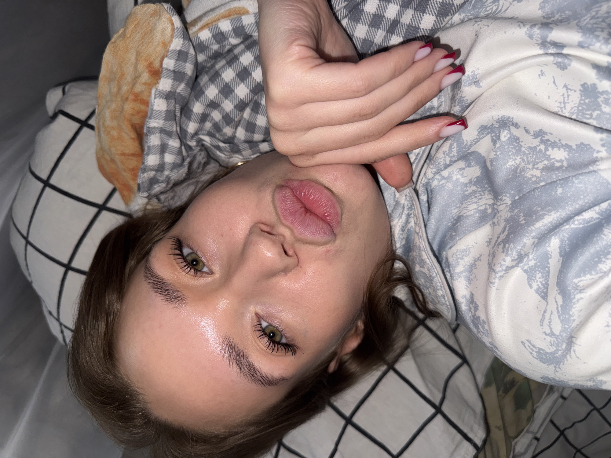A young Caucasian woman with brown hair, green eyes, and full lips is lying down, likely in bed, in what appears to be a residential setting in Chaikovsky, Russia. She is wearing a light blue and white patterned pajama top. Her right hand is resting on her chin and cheek, and her fingernails are painted with a French manicure. She is looking directly at the camera with her mouth slightly puckered. The background consists of pillows and bedding, one of which has a black and white checkered pattern, and another a tan and orange floral pattern. The lighting suggests an indoor setting, possibly with natural light supplemented by artificial light.
