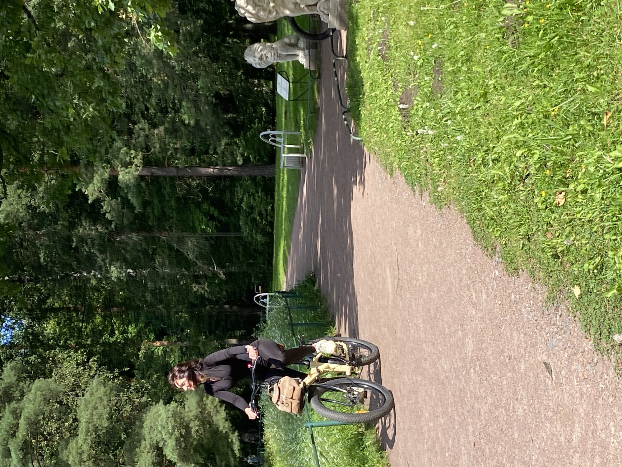A woman is riding a beige bicycle on a paved path in a park. She is wearing a black long-sleeved top and black leggings. A brown bag is attached to the front of her bicycle. The path is bordered by green grass and trees. In the background, there are two stone lion statues and a white bench. The scene appears to be in a public park, likely in the city of Pavlovsk, Russia, which is known for its historical parks and gardens. The lighting suggests it is daytime, with sunlight filtering through the trees.