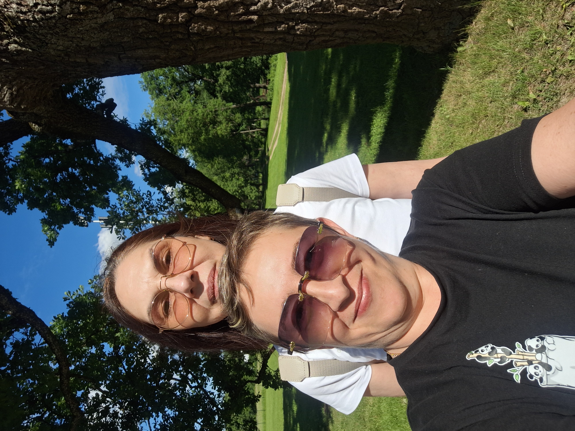 Two women are taking a selfie outdoors. The woman in the foreground is wearing a black t-shirt with a panda graphic and sunglasses. The woman behind her is wearing a white t-shirt and pink-tinted glasses. They are standing in a park with green grass, trees, and a path in the background. A large tree trunk is on the right side of the frame. The sky is blue with some white clouds. The overall setting appears to be a sunny day in a park within the city of Gatchina, Russia.