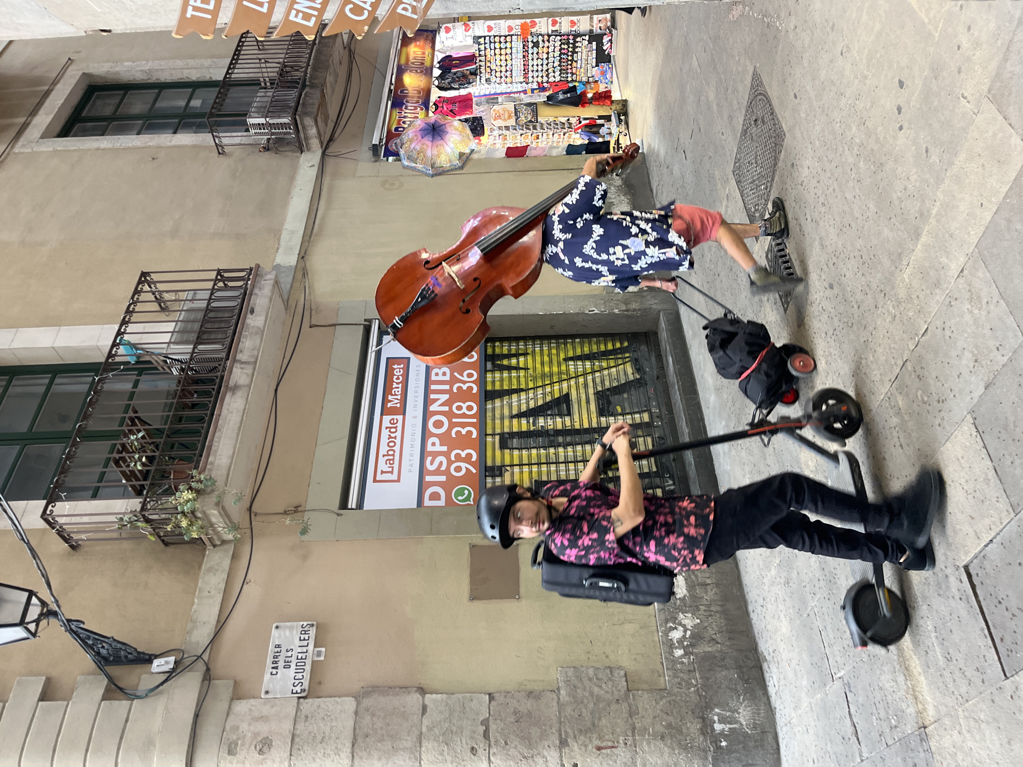 This image captures a vibrant street scene in Barcelona, Spain, featuring two individuals engaged in what appears to be a street performance or daily activity. The setting is a narrow, old street lined with stone buildings and storefronts, typical of a historic city center.
In the mid-ground, a person plays a large cello, its rich brown wood contrasting with their blue and white patterned short-sleeved shirt and orange shorts. The cello is positioned on the pavement, and the person is focused on playing the instrument. Nearby, a black bag on wheels is visible, possibly holding equipment. In the foreground, another individual stands holding an electric scooter. They wear a black helmet, a black and pink patterned shirt, and black trousers. Their posture suggests they are either waiting, observing, or preparing to move.
The background reveals a bustling urban environment. On the right, a shop or stall displays various souvenirs, including t-shirts and small decorative items, with visible text "Batiga Barcelona" (though OCR suggested "Satiga Belong") and "VODKA", "COCK" likely referring to drinks or cocktails. The buildings feature traditional wrought-iron balconies. On the left wall, a street sign clearly reads "CARRER DELS ESCUDELLERS," identifying the location. Below this, a commercial sign for "Laborde Marcet, PATRIMONIO & INVERSIONES" with the text "DISPONIB" (likely "Disponible" meaning available) and a phone number "93 318 36" is visible. The scene is illuminated by daylight, suggesting a clear day, and exudes a lively, active atmosphere.